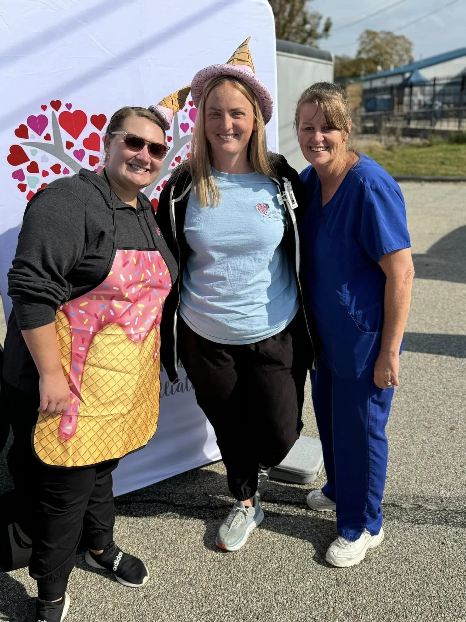 Three women smiling outdoors, one wearing a pink hat with cone shapes and a pink ice cream cone apron, and another in blue medical scrubs, standing in front of a backdrop with red and pink hearts.