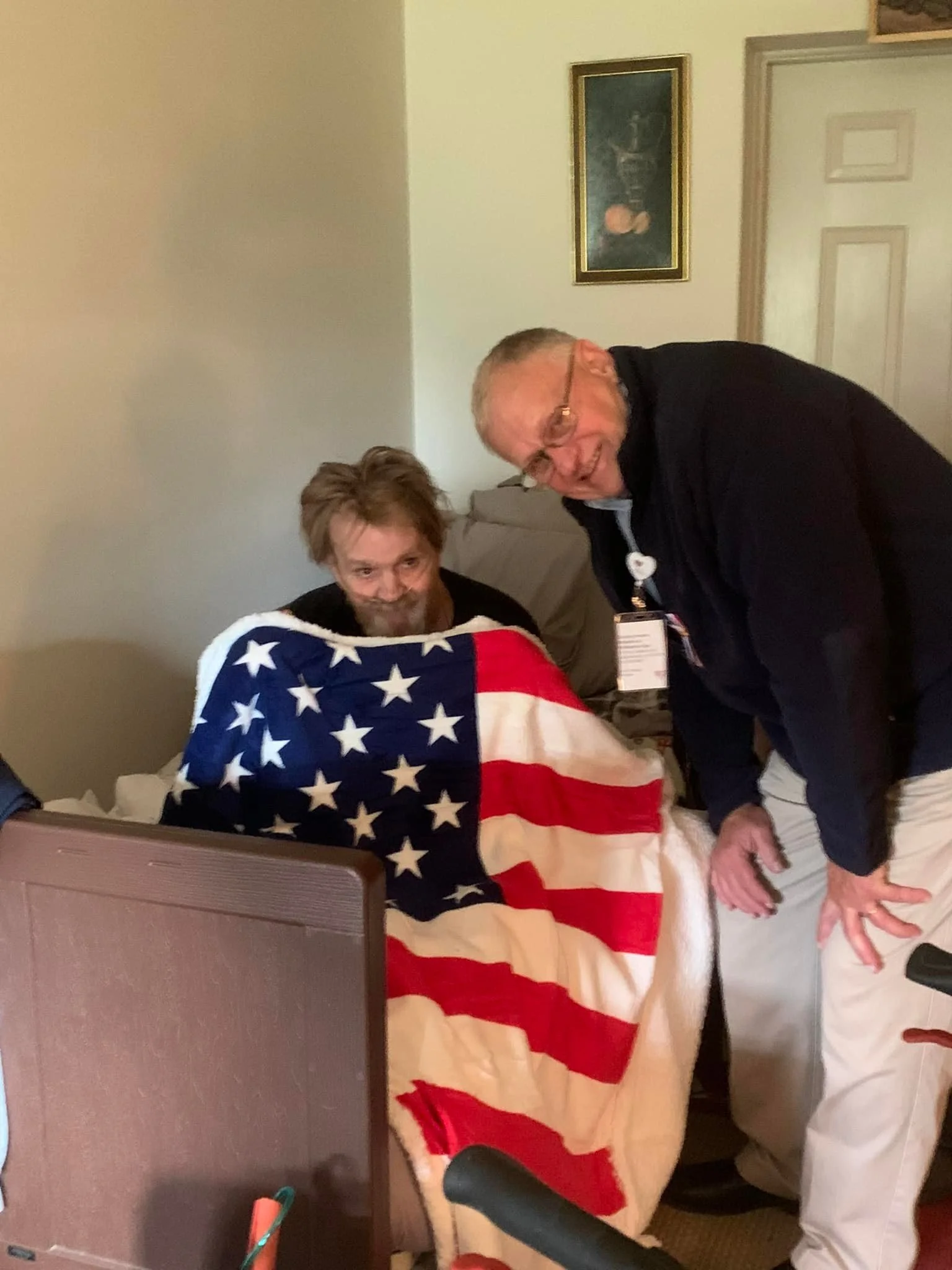 Two men, one sitting on a bed with an American flag blanket, and another standing next to him smiling. The man on the bed has a beard and short hair, and the standing man has glasses and gray hair.