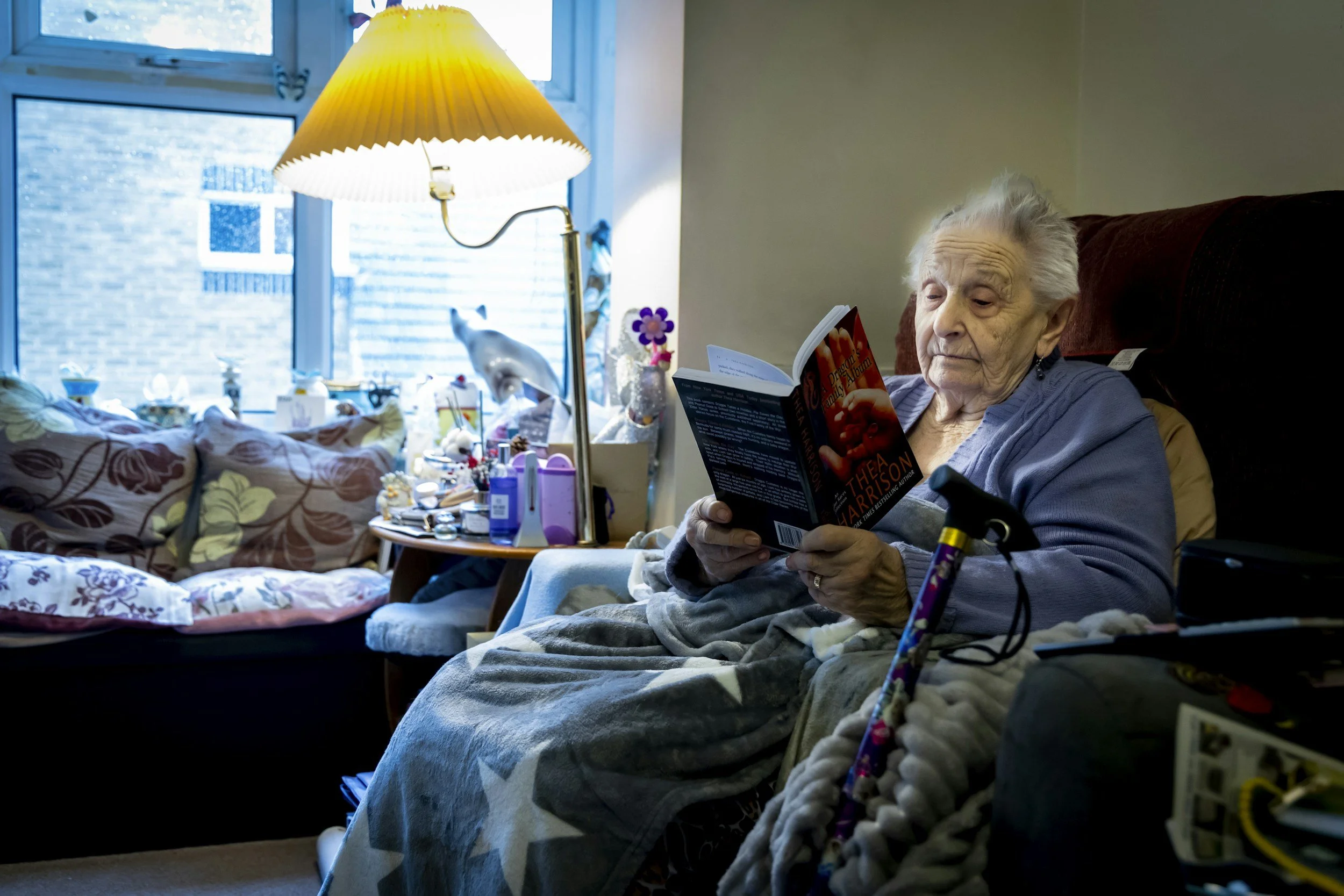 An elderly woman sitting in a wheelchair, reading a book in a cozy living room with a window, a table with various items, and a yellow lamp.