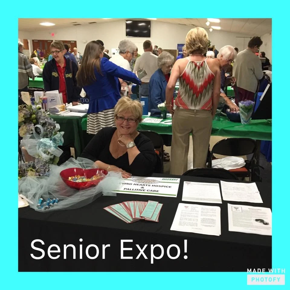 A woman sitting at a table with the sign 'Living Hearts Hospice & Palliative Care' at a senior expo. The table has brochures, pamphlets, and a bowl of candies. There are many seniors and older women standing in the background at the event.