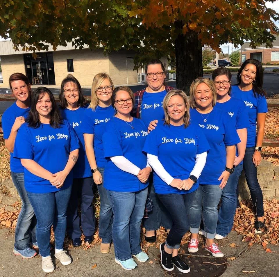 Group of ten women standing outdoors under a tree, wearing matching blue t-shirts with 'Love for Libby' printed on them, posing for a photo with fallen leaves on the ground.