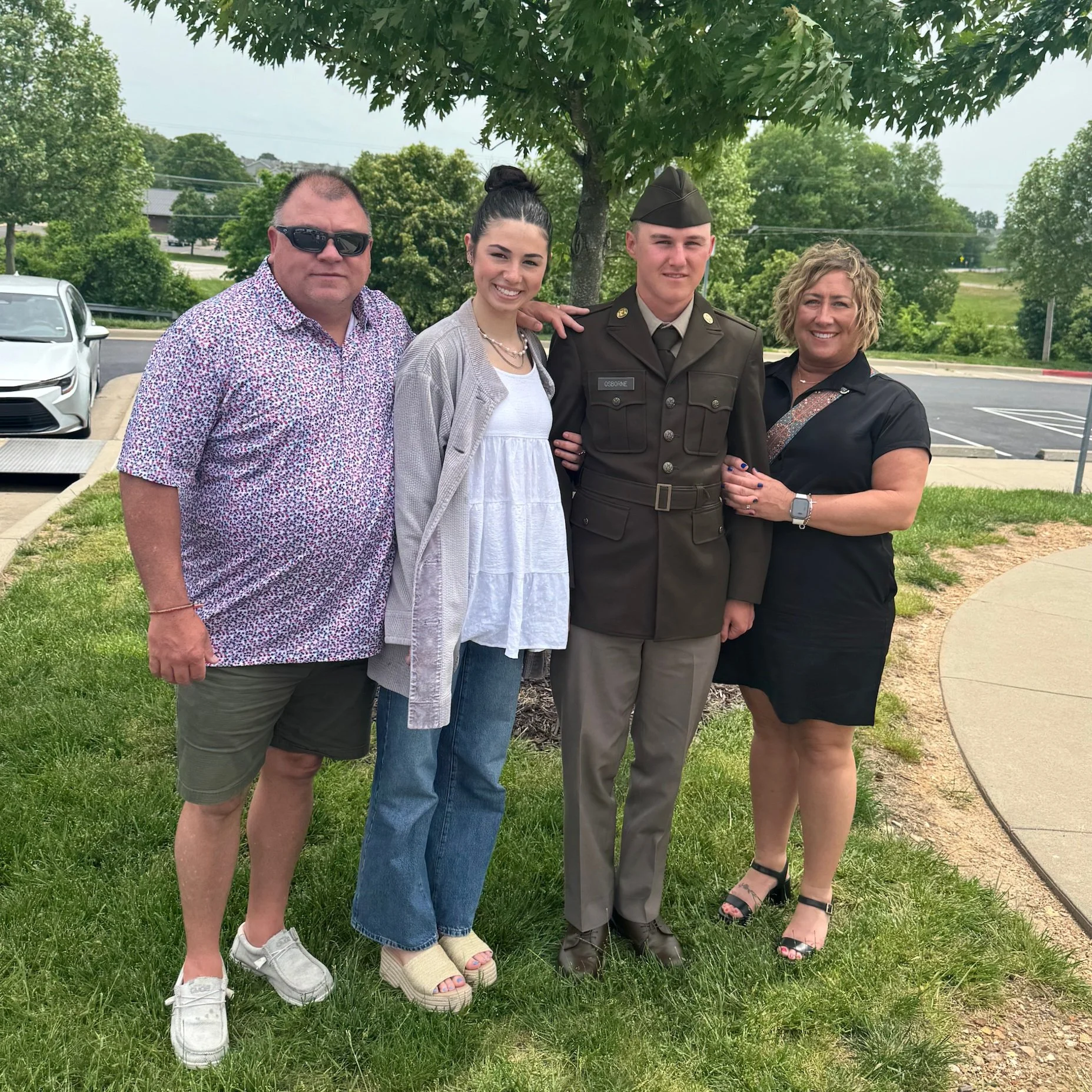 A group of four people standing outdoors on a grassy area, posing for a photograph. The group includes a man wearing sunglasses and a short-sleeved patterned shirt, a young woman with dark hair in a bun wearing a white dress and a light coat, a young man in a military uniform, and a woman in a black dress. They are smiling and standing close together in front of a tree, with a parking lot and some greenery in the background.