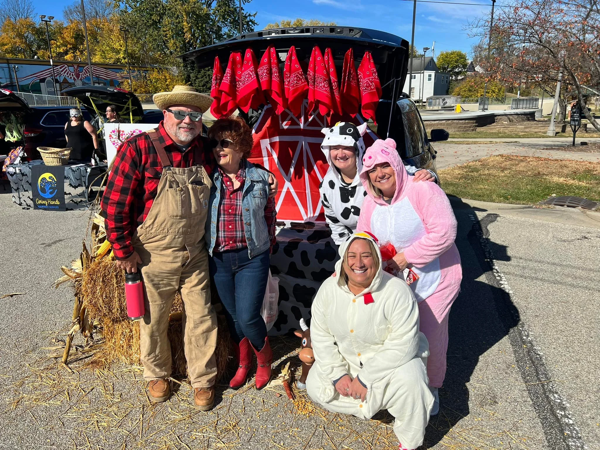 Group of five people dressed in cow, pig, and sheep costumes posing in front of a decorated vehicle with a red barn design during a fall outdoor event. Autumn leaves and trees are visible in the background.