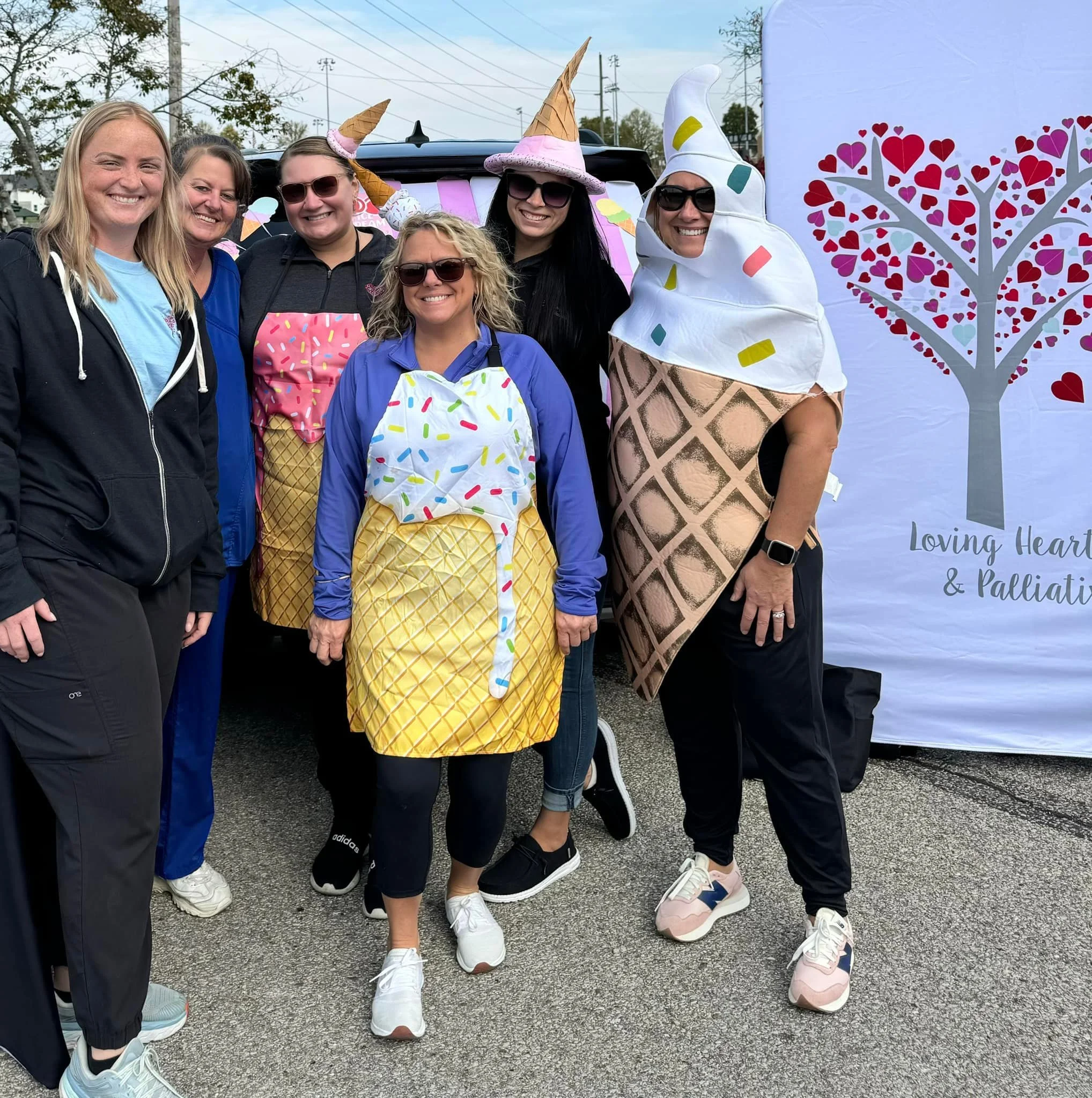 Group of women wearing ice cream cone costumes and casual clothing, standing outdoors near a decorated backdrop with a heart-shaped tree and the words "Loving Heart & Palliative."