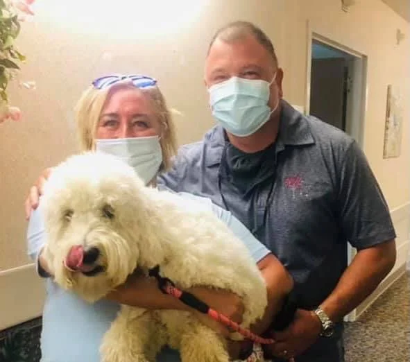 Two people wearing masks holding a large, fluffy white dog on a leash indoors.