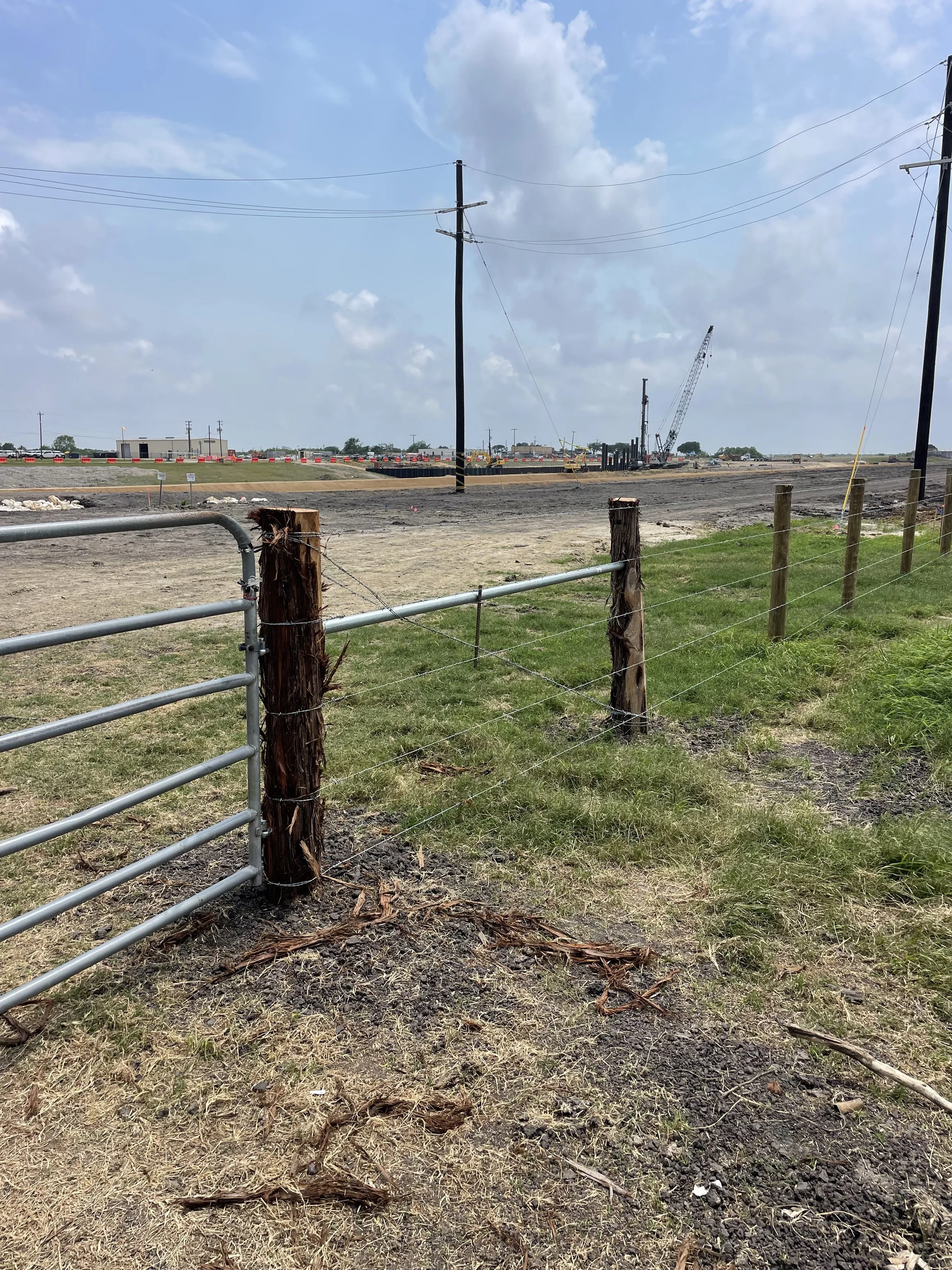 A rural construction site with a new fence, utility poles, and a dirt area with construction equipment in the distance under a partly cloudy sky.