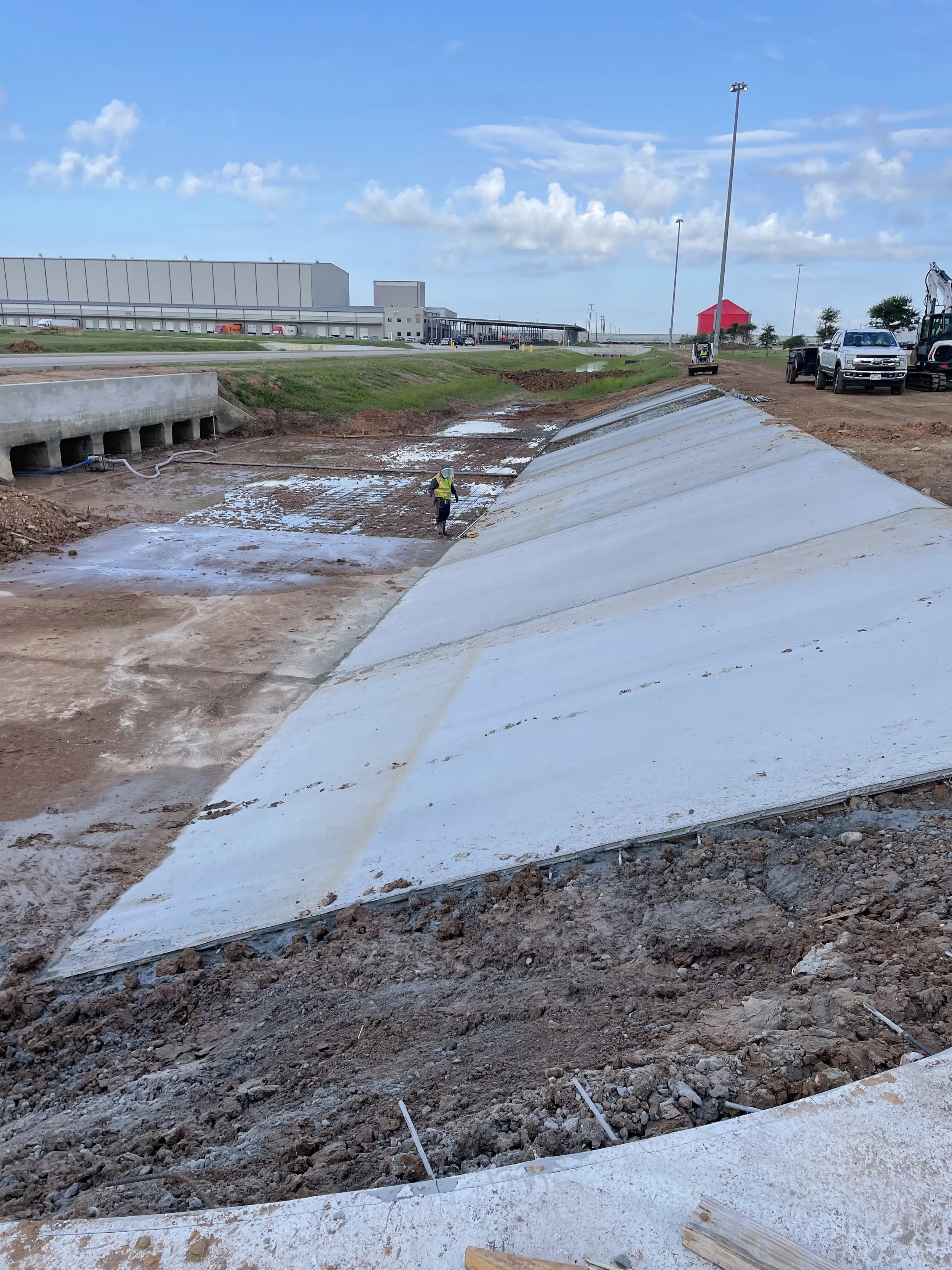 Construction site with a worker in a safety vest and helmet working on a large concrete retaining wall or ramp, construction vehicles and equipment in the background, under a partly cloudy sky.