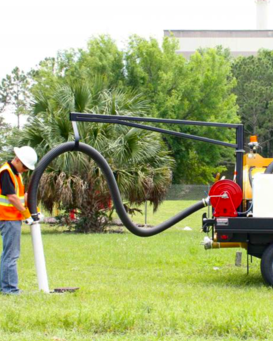Worker in safety vest and helmet operates vacuum machine in a grassy outdoor area with trees.