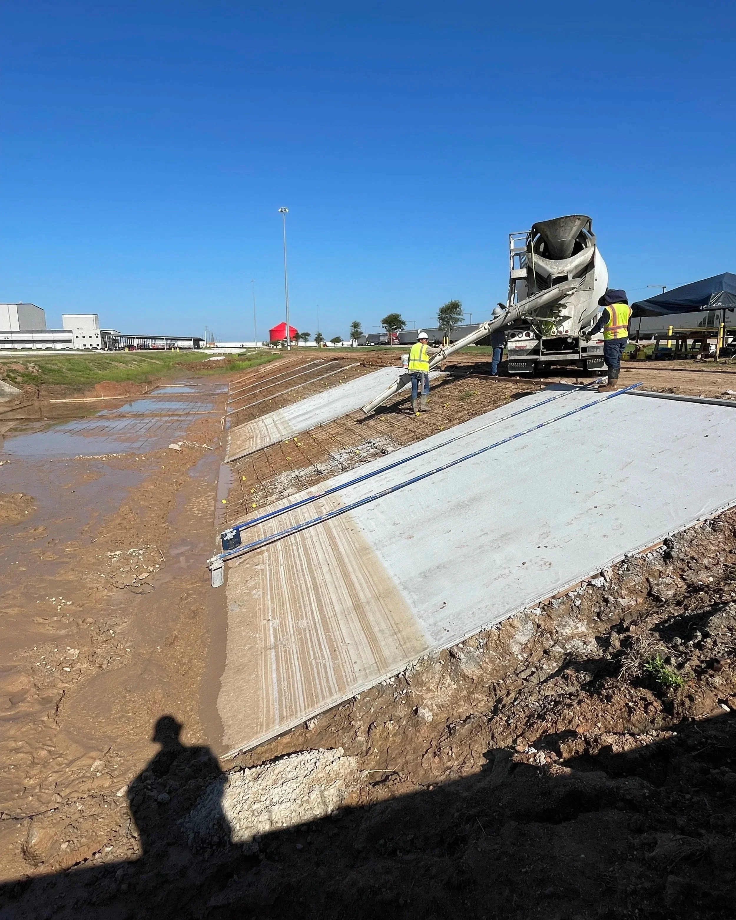 Construction workers pouring concrete on a sloped surface at a construction site with clear blue skies and a few distant trees and buildings.