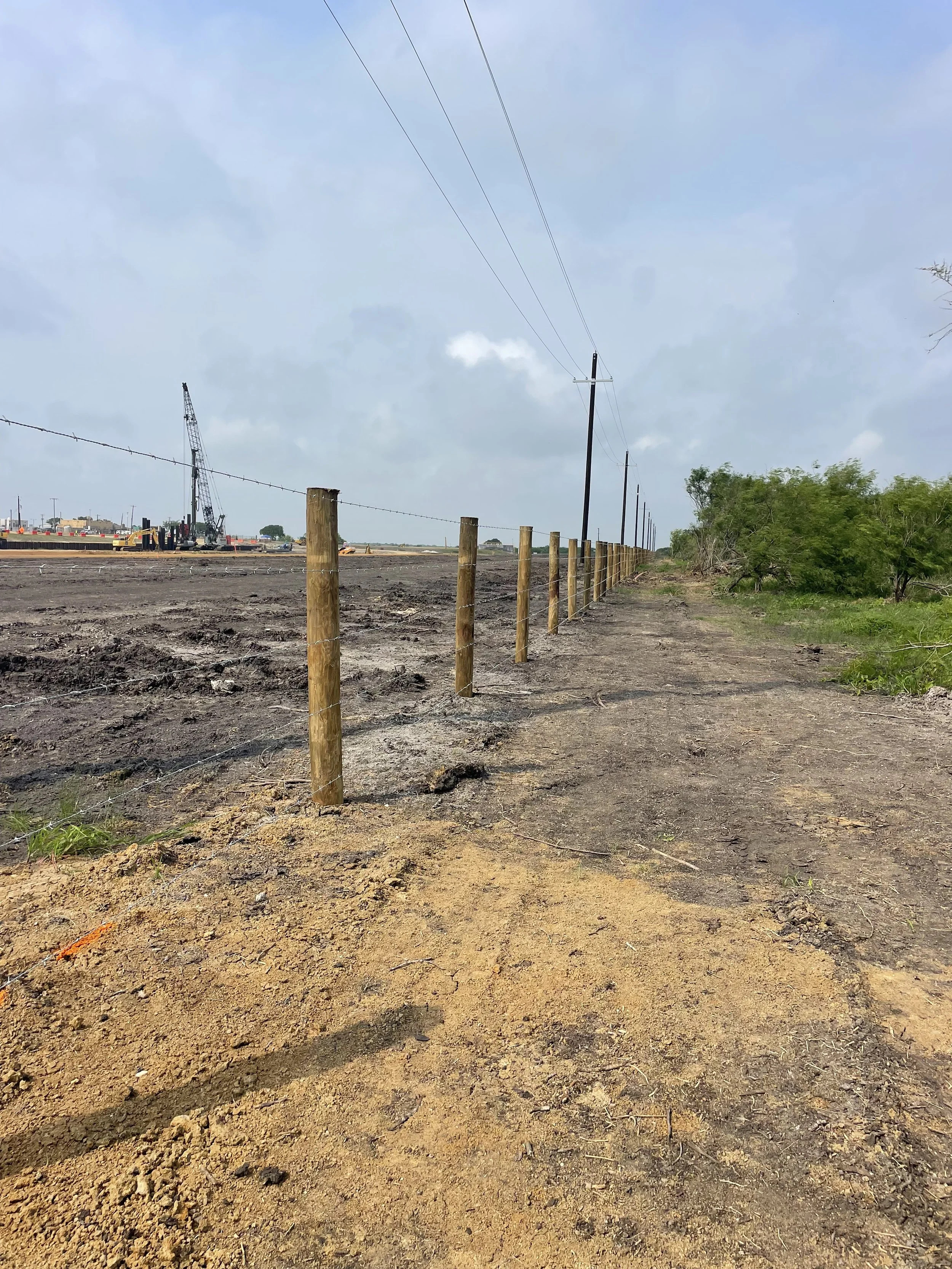 A dirt path runs alongside a fence with wooden posts and wires. To the left, there is a construction site with cranes and equipment in the distance. To the right, there are green trees and bushes. Overhead, power lines run parallel to the path, under