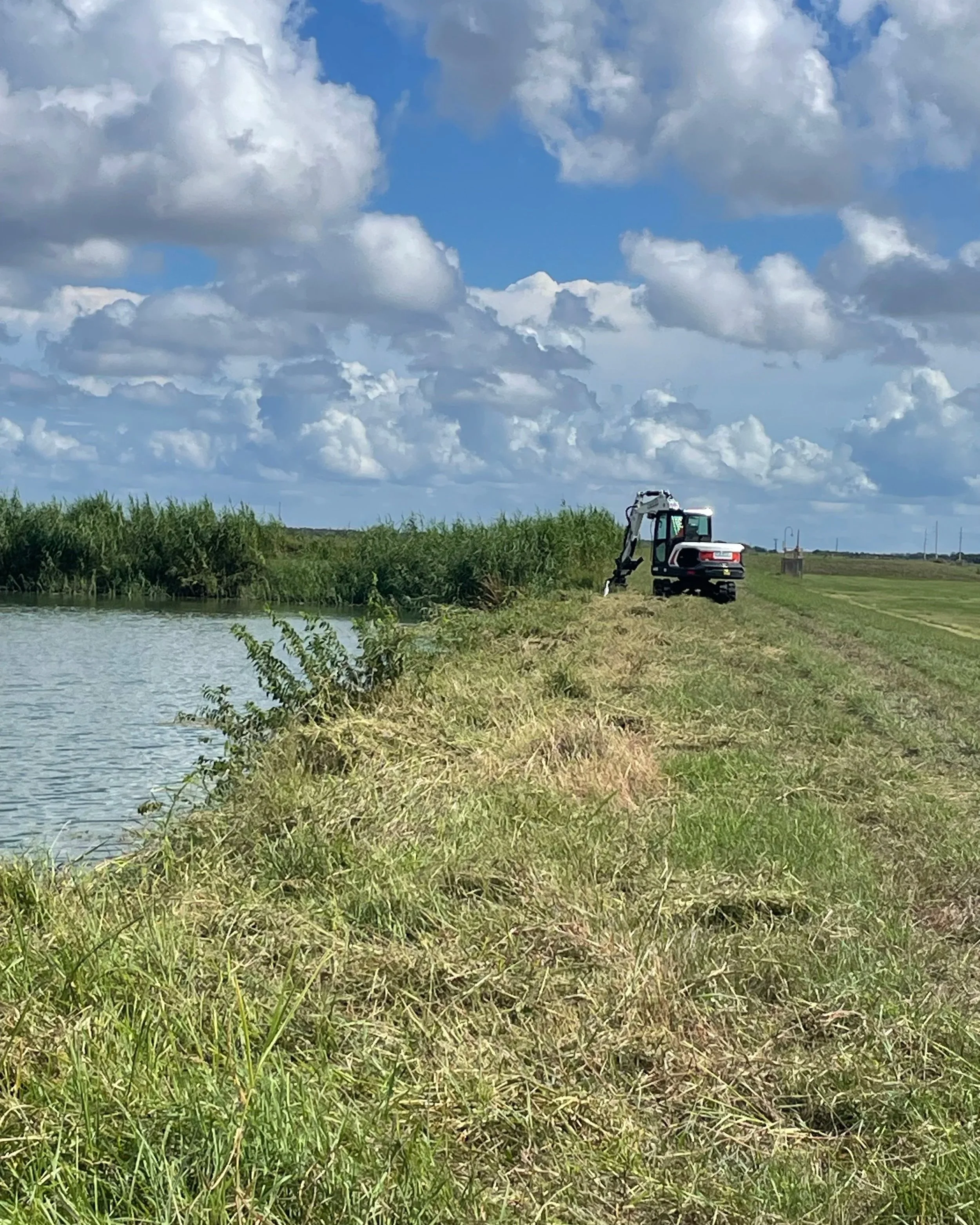 A small excavator working beside a body of water on a grassy bank under a partly cloudy sky.