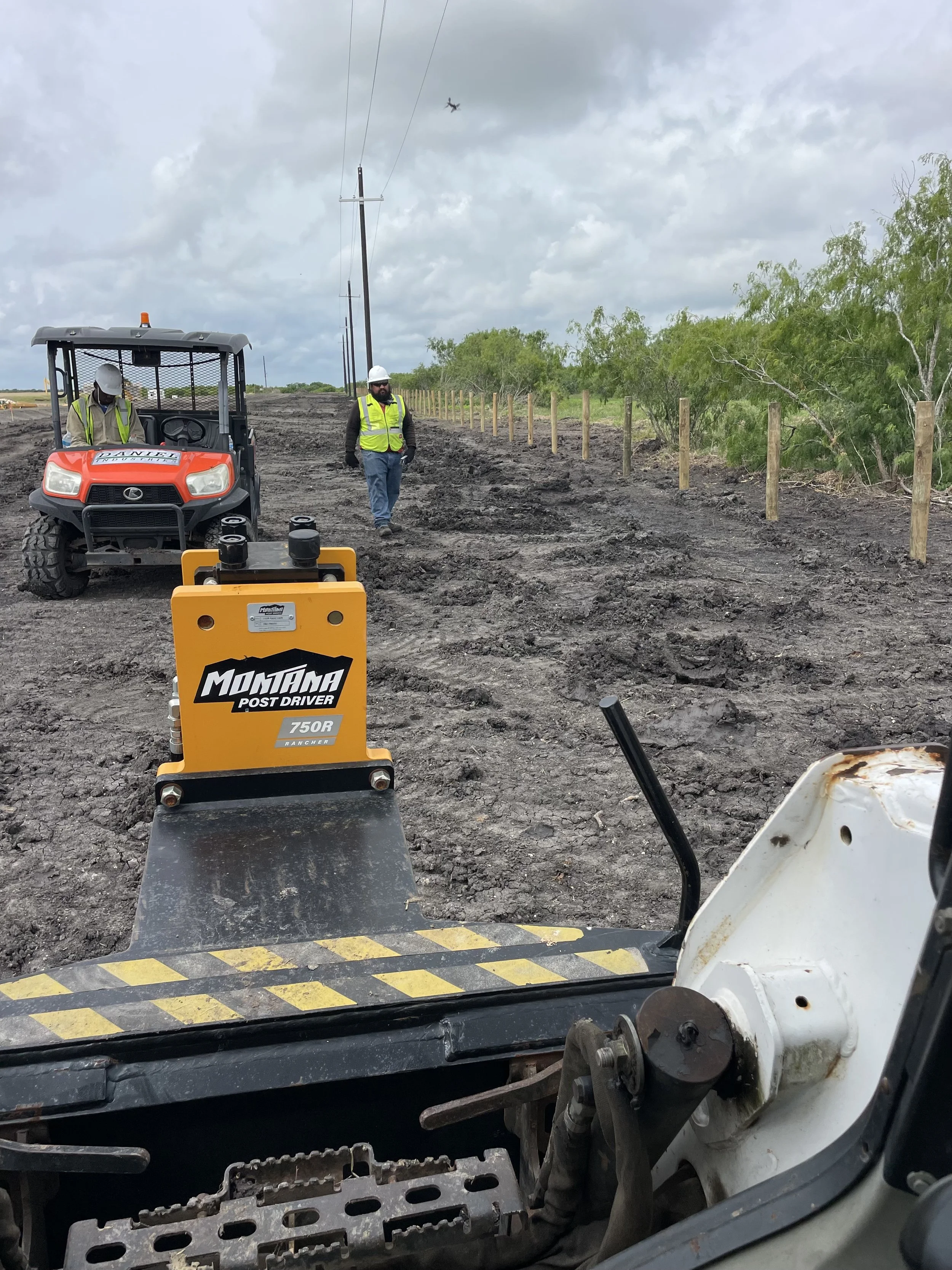Workers in safety vests and helmets working on a construction site with tools, a utility vehicle, and wooden posts, under a cloudy sky.