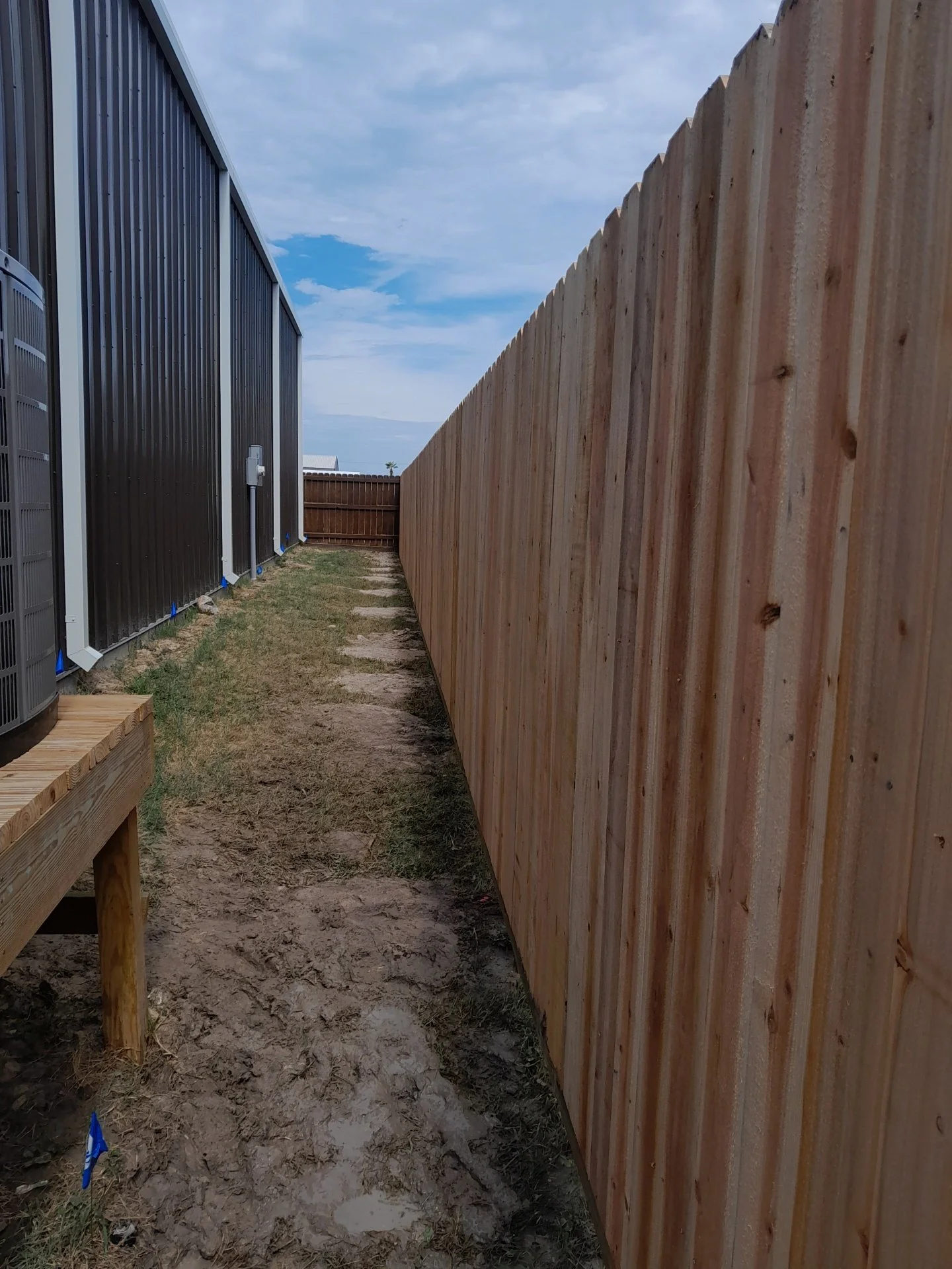 Side yard with a wooden fence on the right and outdoor air conditioning units on the left, with a narrow dirt pathway in between.