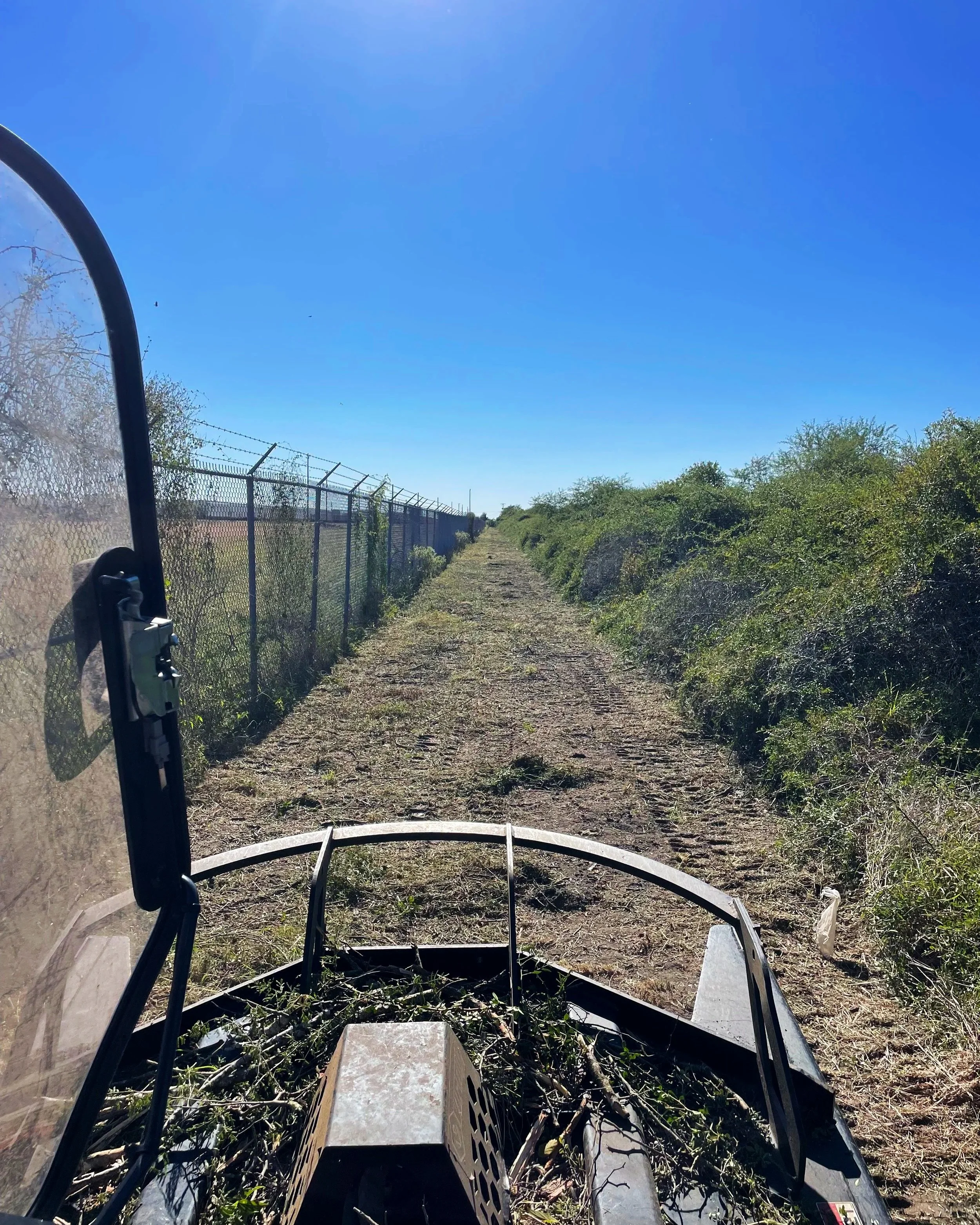 View from a vehicle, showing a dirt trail with tire tracks, a chain-link fence on the left, green bushes on the right, and a clear blue sky.