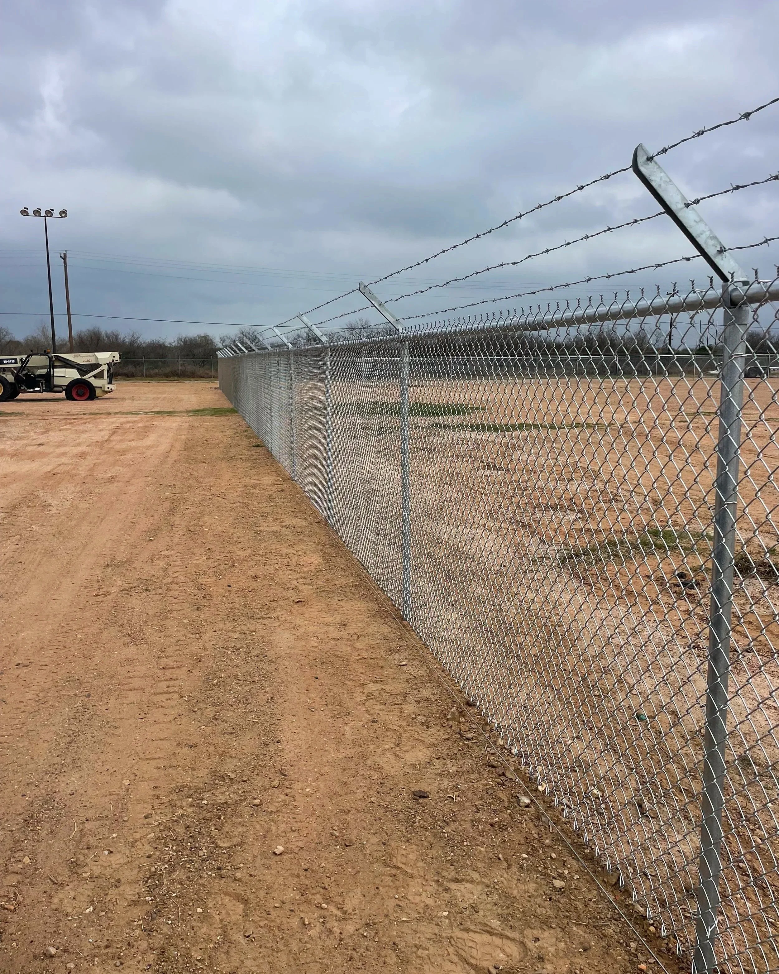 A chain-link fence topped with barbed wire runs along a dirt area under a cloudy sky, with vehicle and light poles in the background.