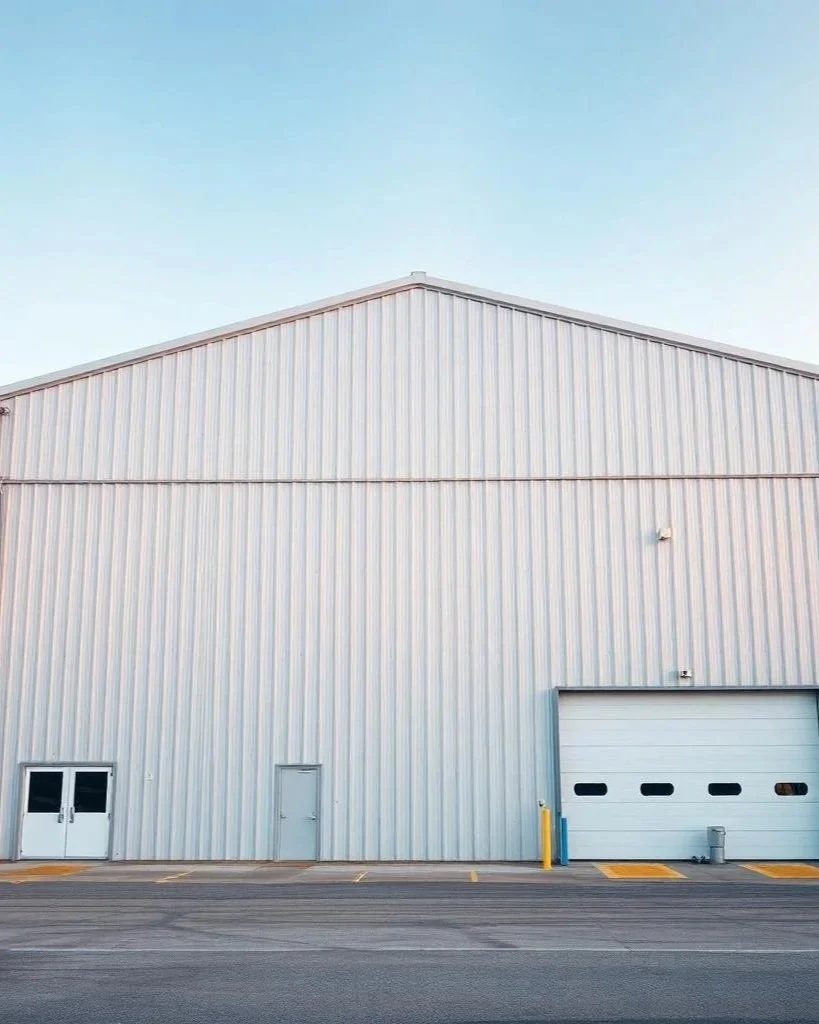 Large metal warehouse building with a roller door, a small pedestrian door, and double doors on a paved lot under a clear blue sky.
