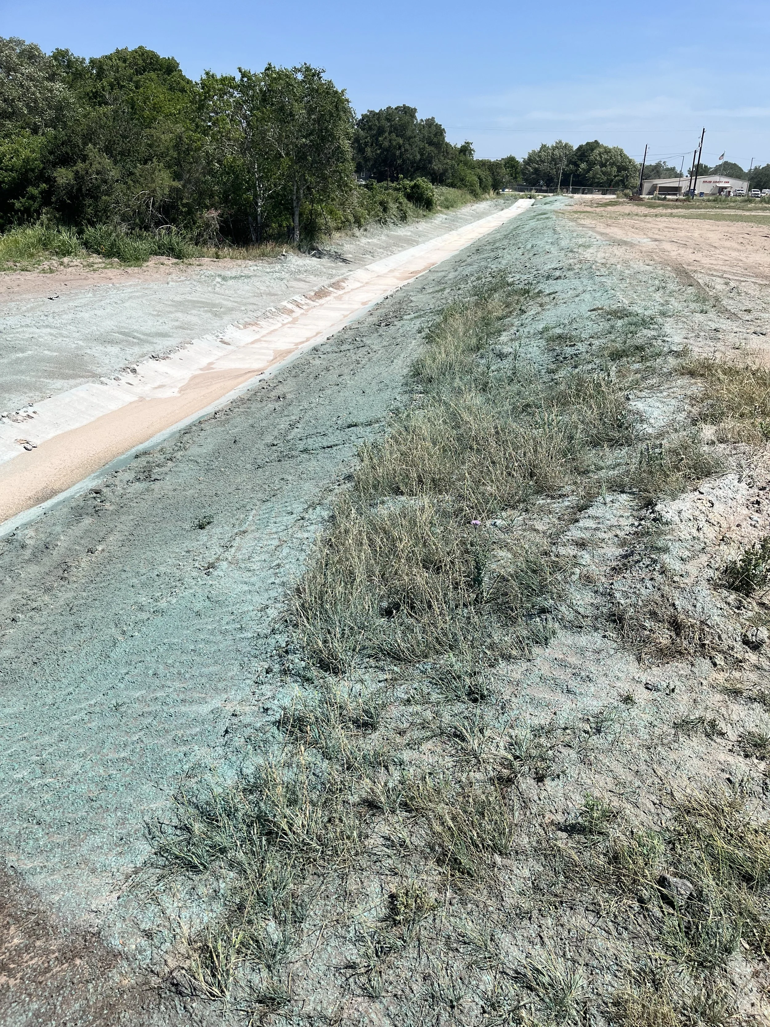 A drainage ditch covered with a layer of pink and white sand, bordered by grass and small plants, with trees in the background and a clear blue sky.
