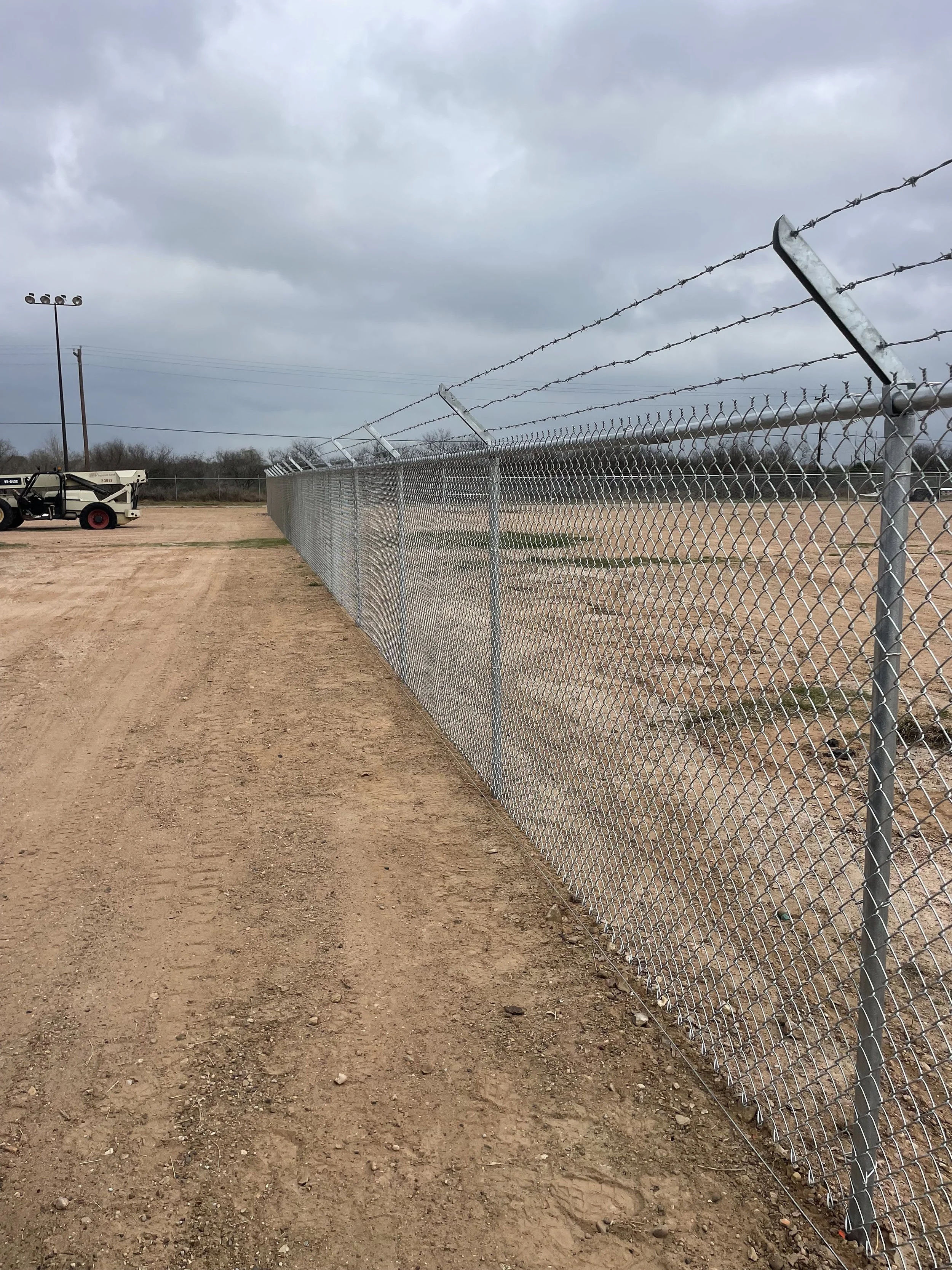 A chain-link fence topped with barbed wire running parallel over a dirt field on a cloudy day, with a utility vehicle in the background.