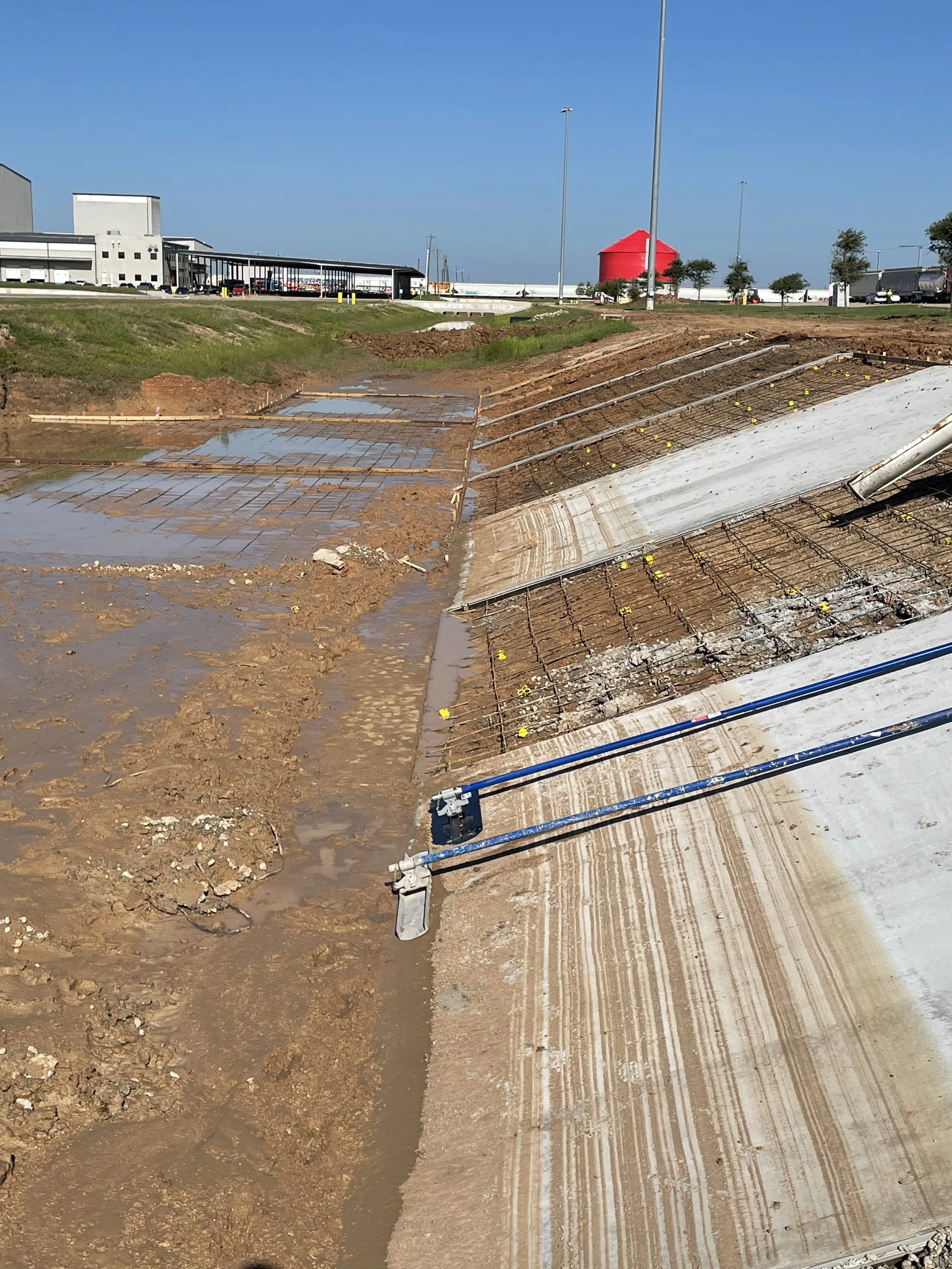Construction site with sloped concrete and metal rebar, plumbing pipes, and muddy ground, with industrial buildings and a red water tower in the background.