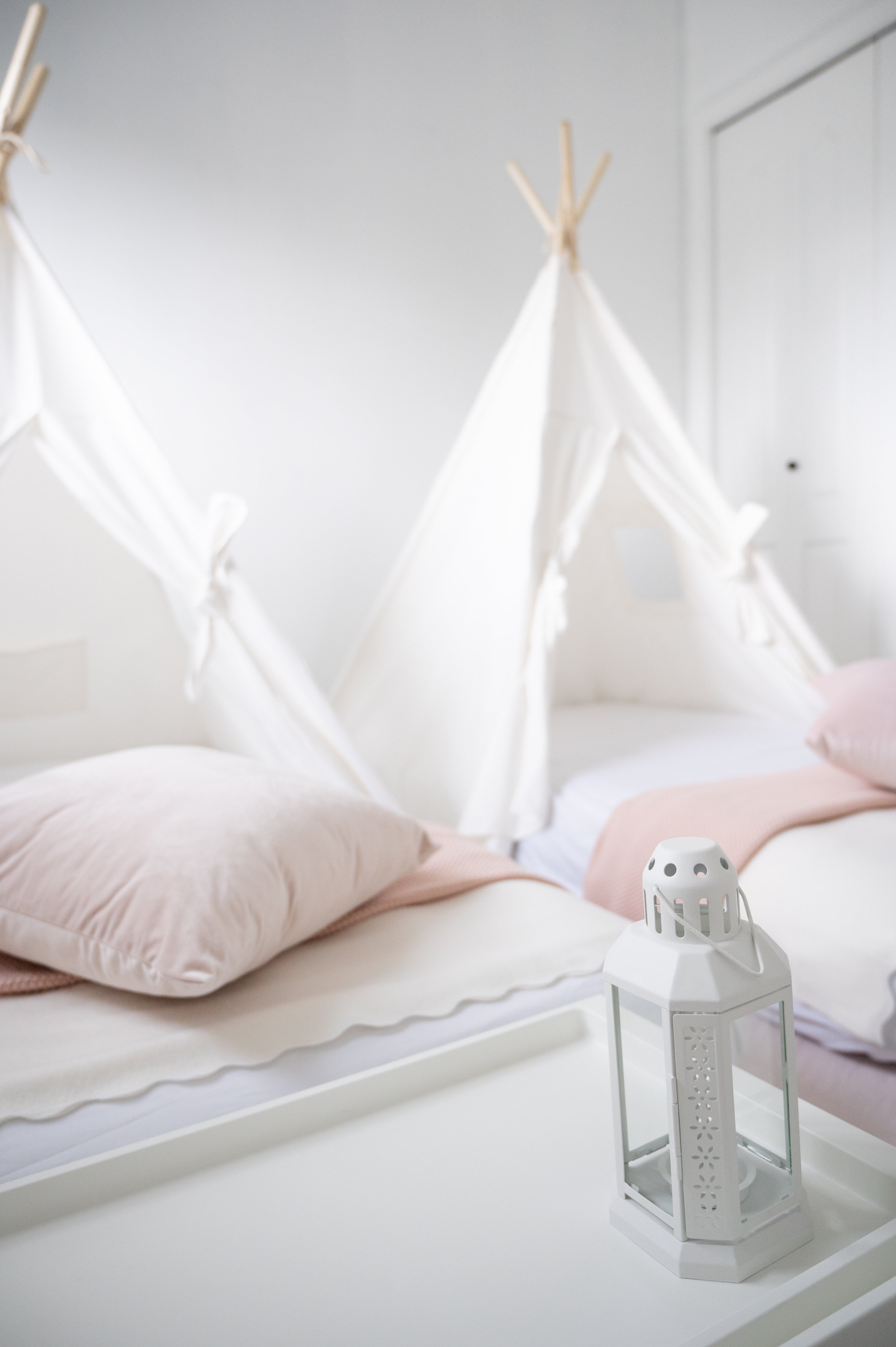 A bedroom with two beds with pale pink pillows and blankets, white canopy beds with knotted fabric, and a white lantern on a tray in the foreground.