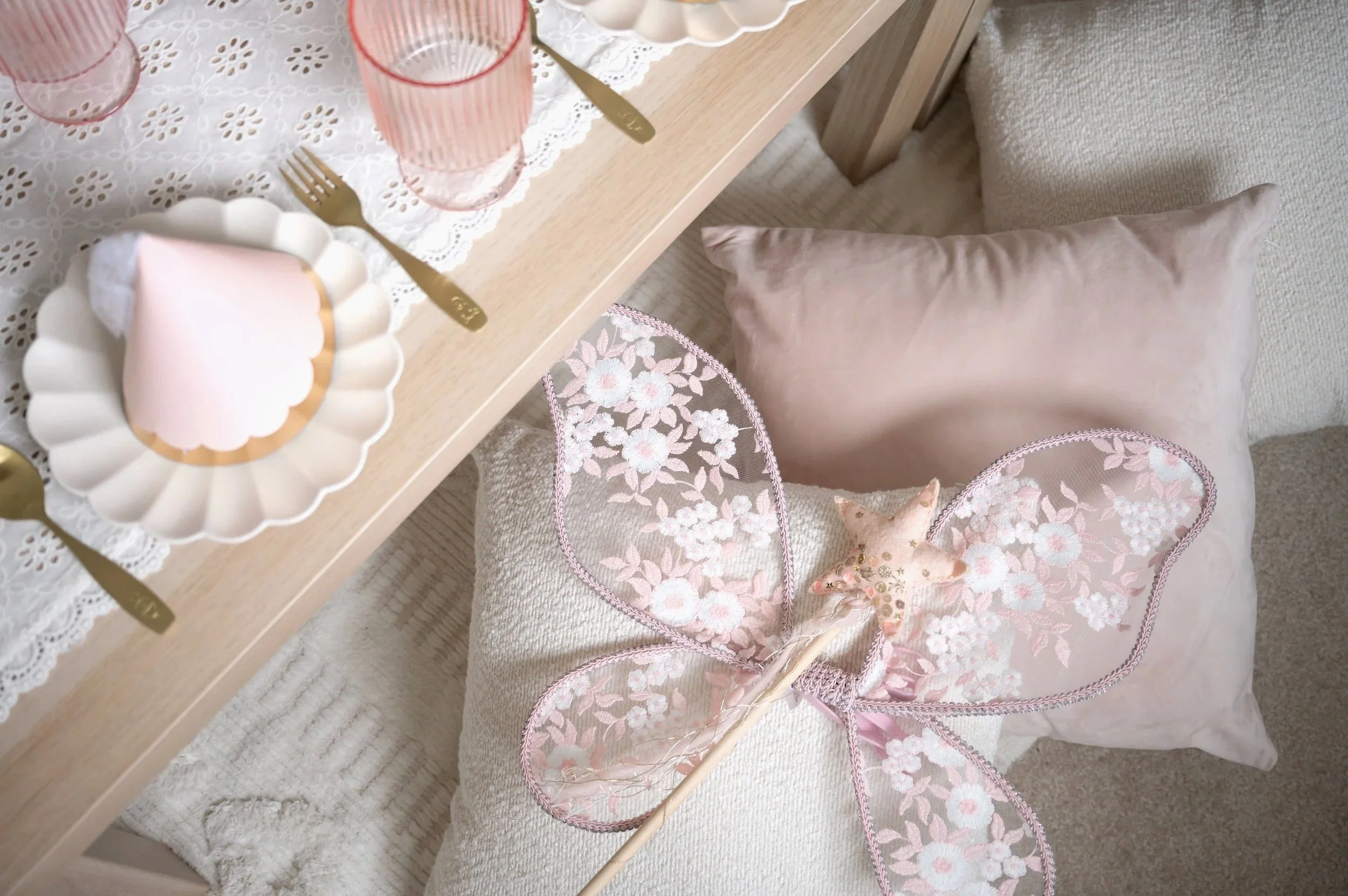 A three-tiered white dessert stand holding pink and beige macarons, cupcakes with white frosting, and small desserts on a lace tablecloth, with additional dessert plates and cupcakes in the background.
