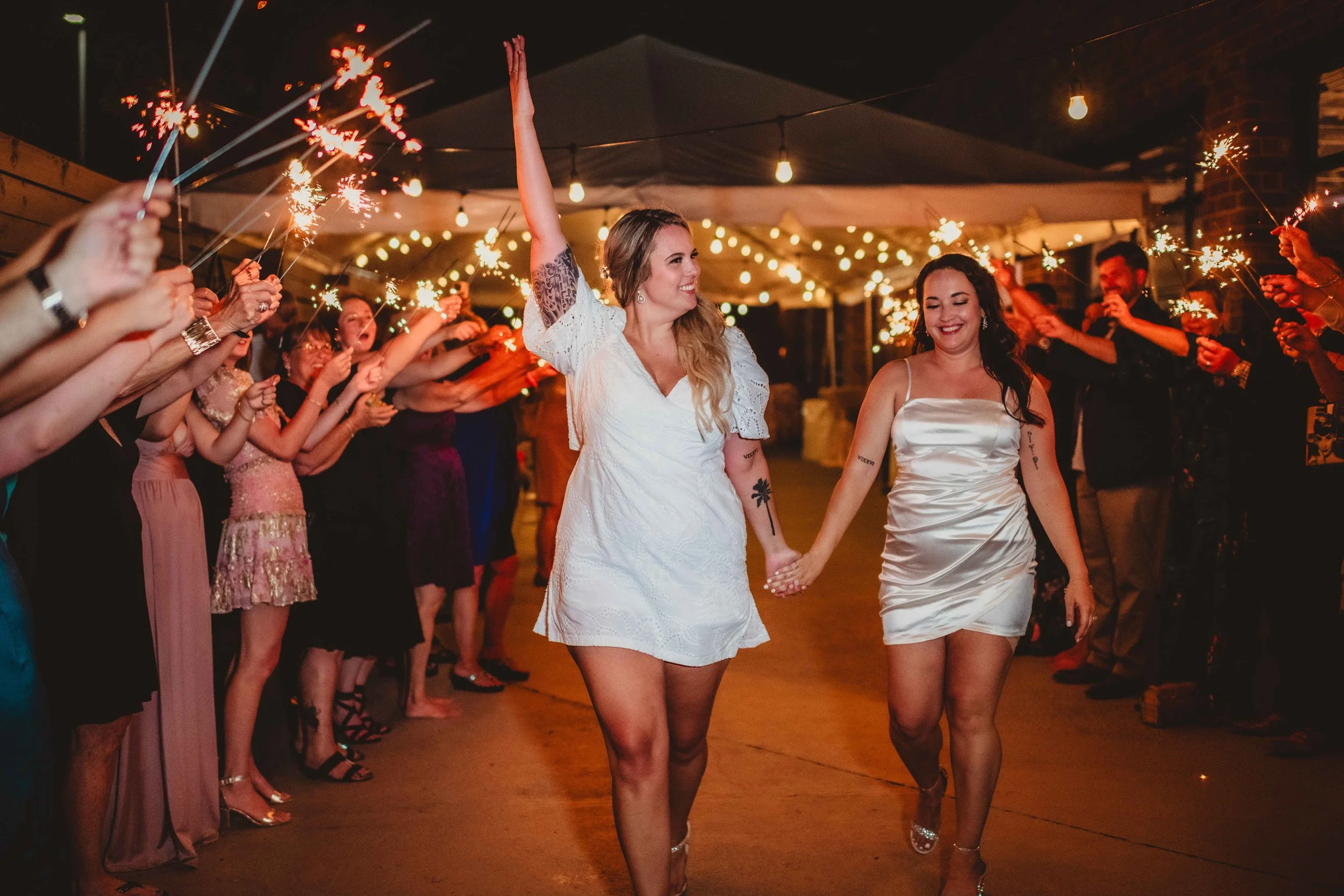 Two women in elegant dresses exit a celebration, surrounded by guests holding sparklers, under string lights.