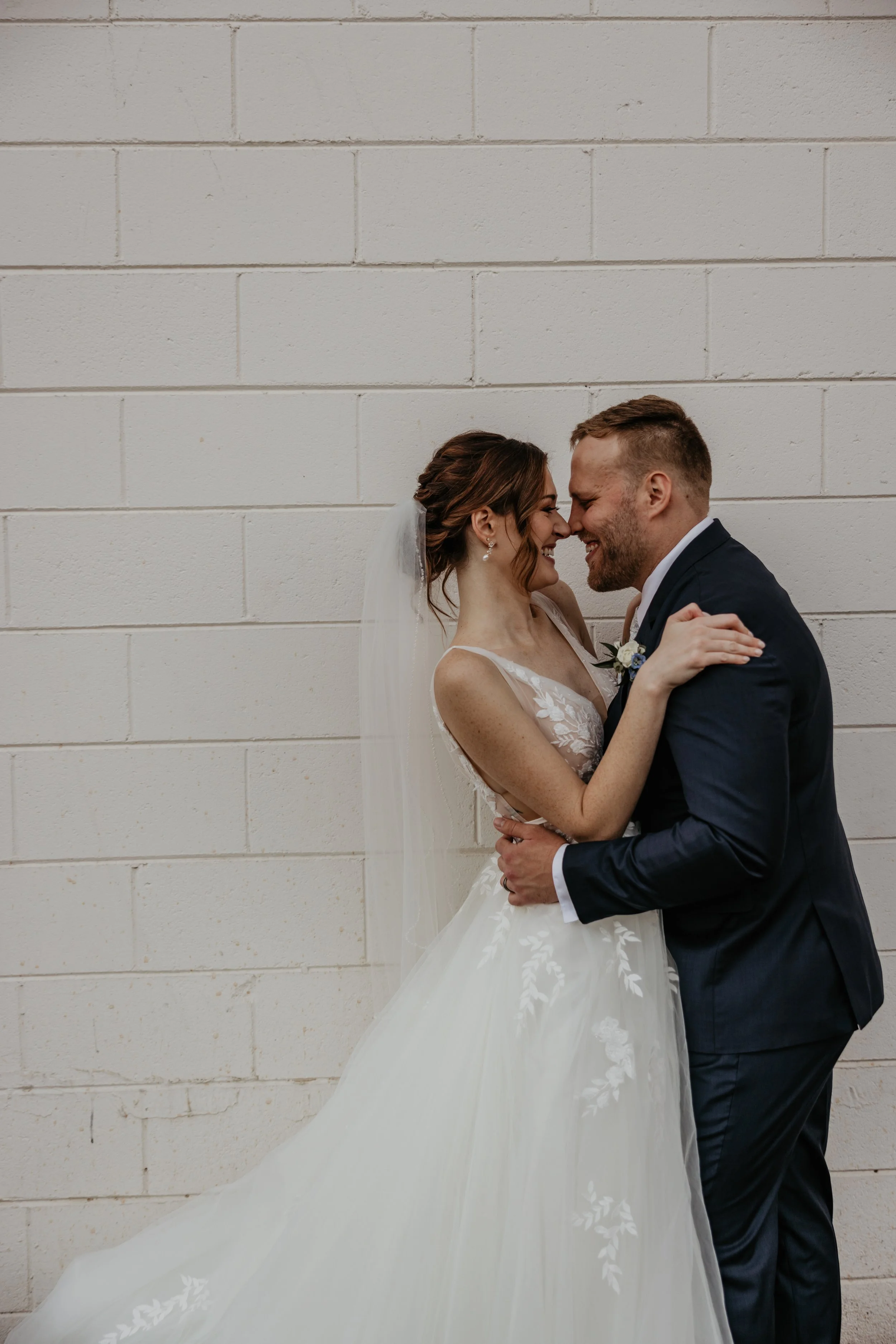 A bride and groom embrace happily in front of a white brick wall. The bride wears a white gown and veil, while the groom is in a dark suit.