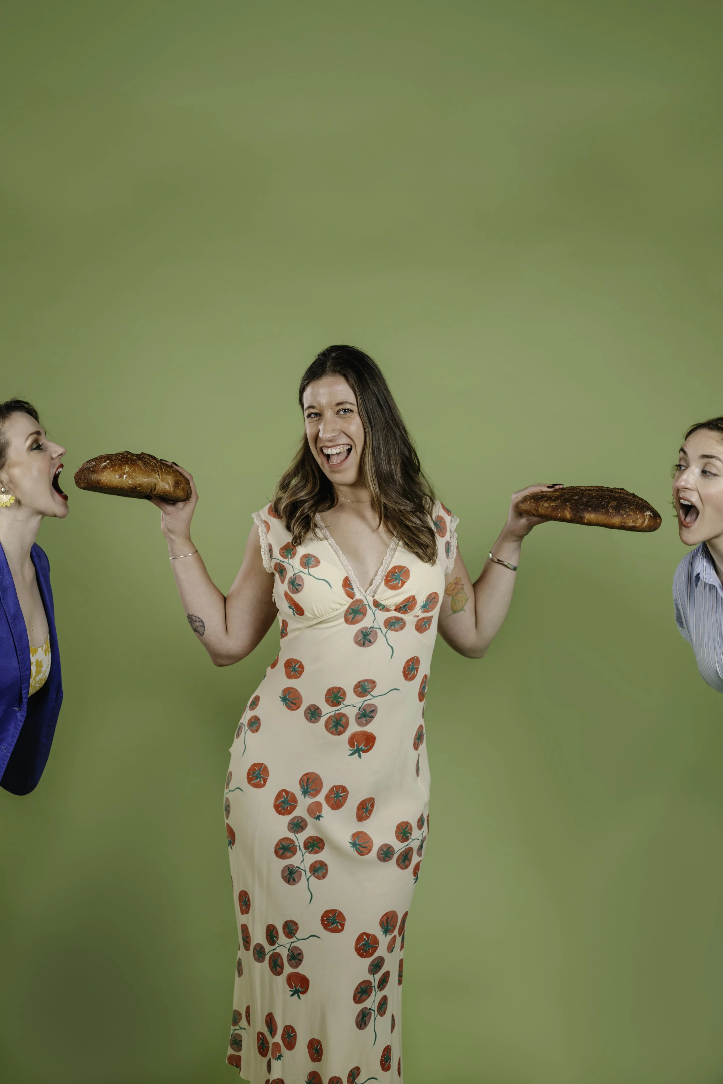 Three women, one in the center wearing a long dress with a tomato print, holding two loaves of bread, smiling and engaging with the other two women on each side, against a green background.