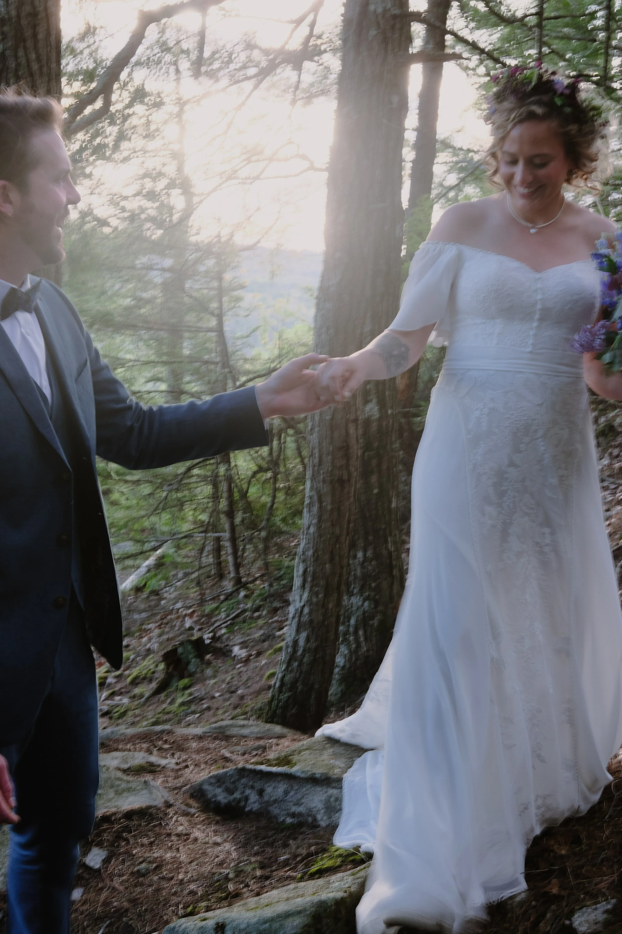 A couple holding hands in a forest, the woman wearing a white wedding dress with floral crown, and the man dressed in a dark suit, both smiling.