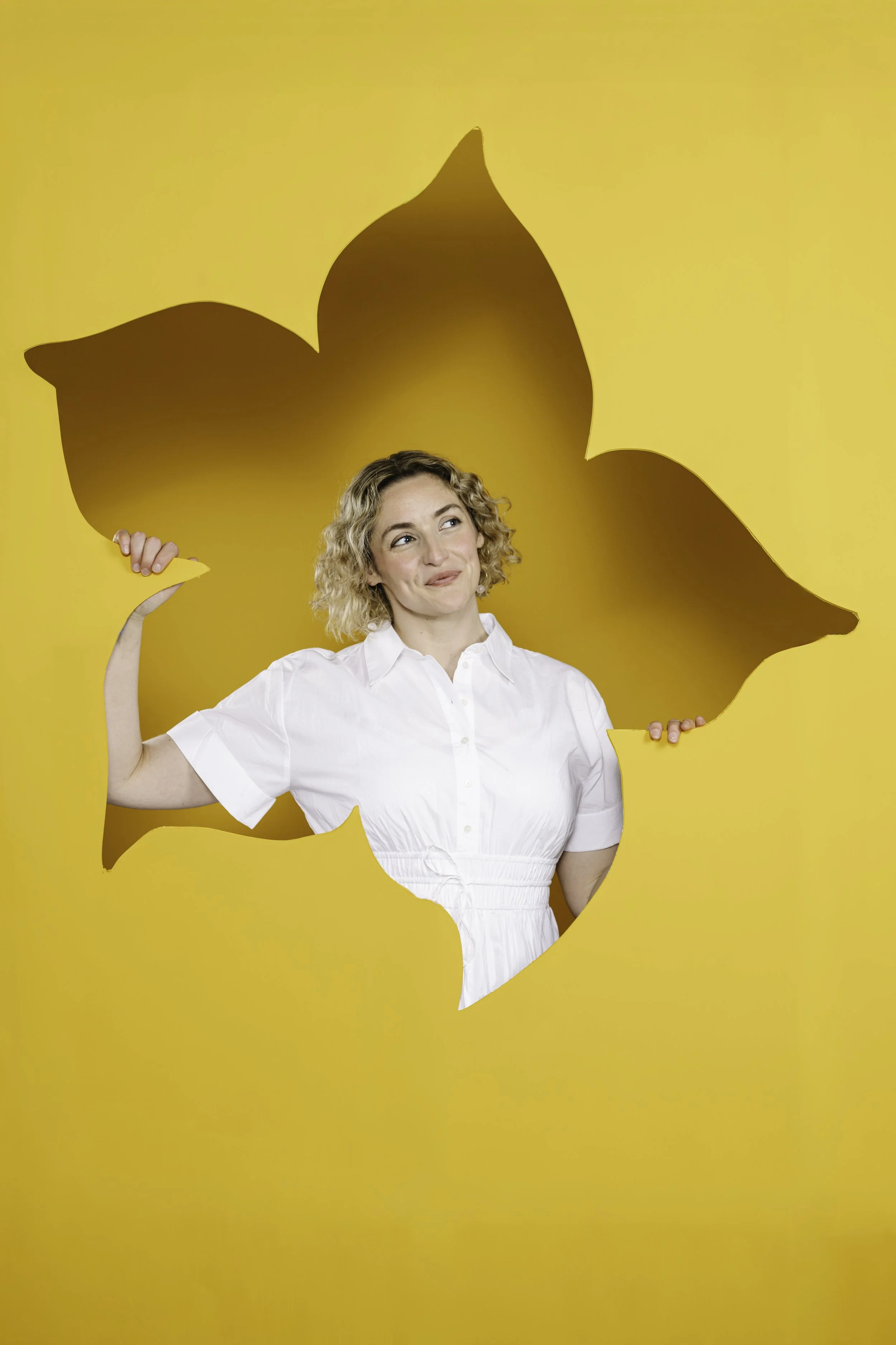 A young woman with blond curly hair in a white dress holds a torn piece of yellow paper with a silhouette of a cat in front of a yellow background, creating a playful illusion.