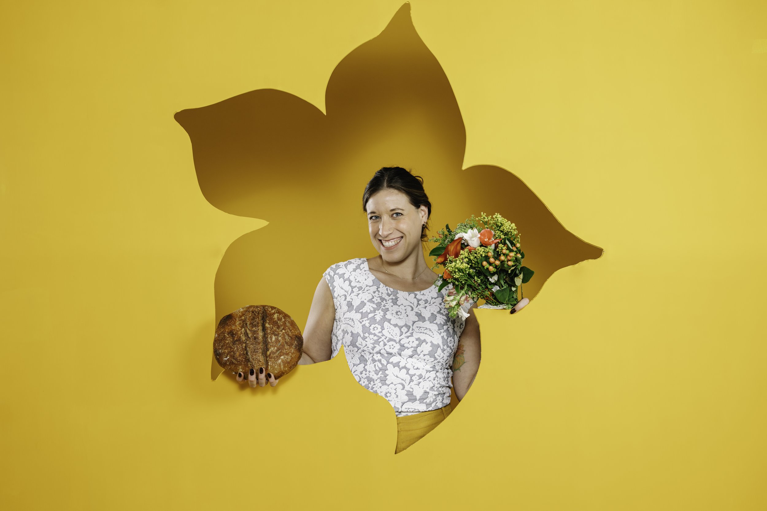 A woman smiling and holding a bouquet of flowers and a loaf of bread against a yellow background with a large leaf-shaped shadow.