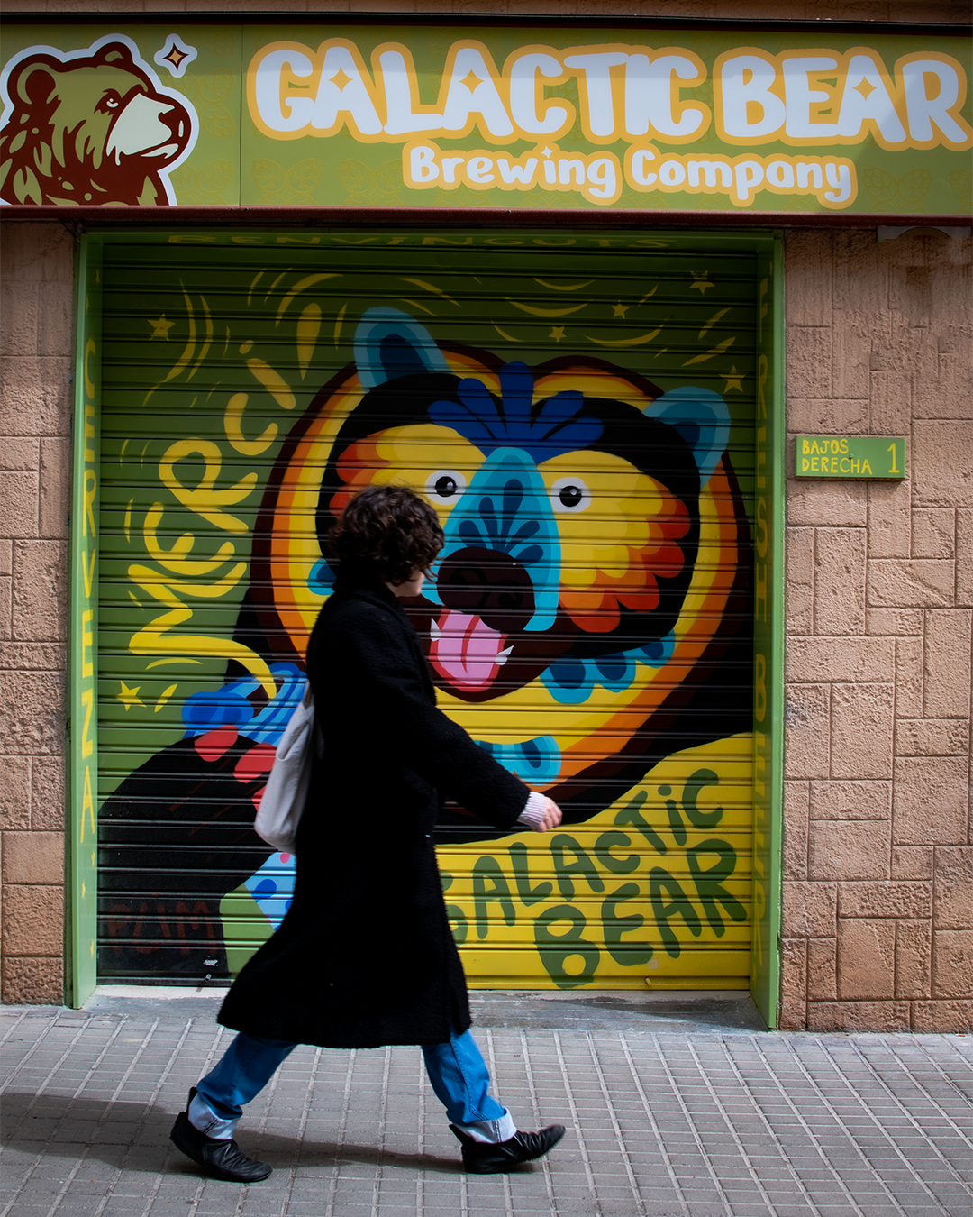 Persona caminando frente a un mural colorido en la puerta de un local llamado Galactic Bear Brewing Company, con un oso de colores en el mural.