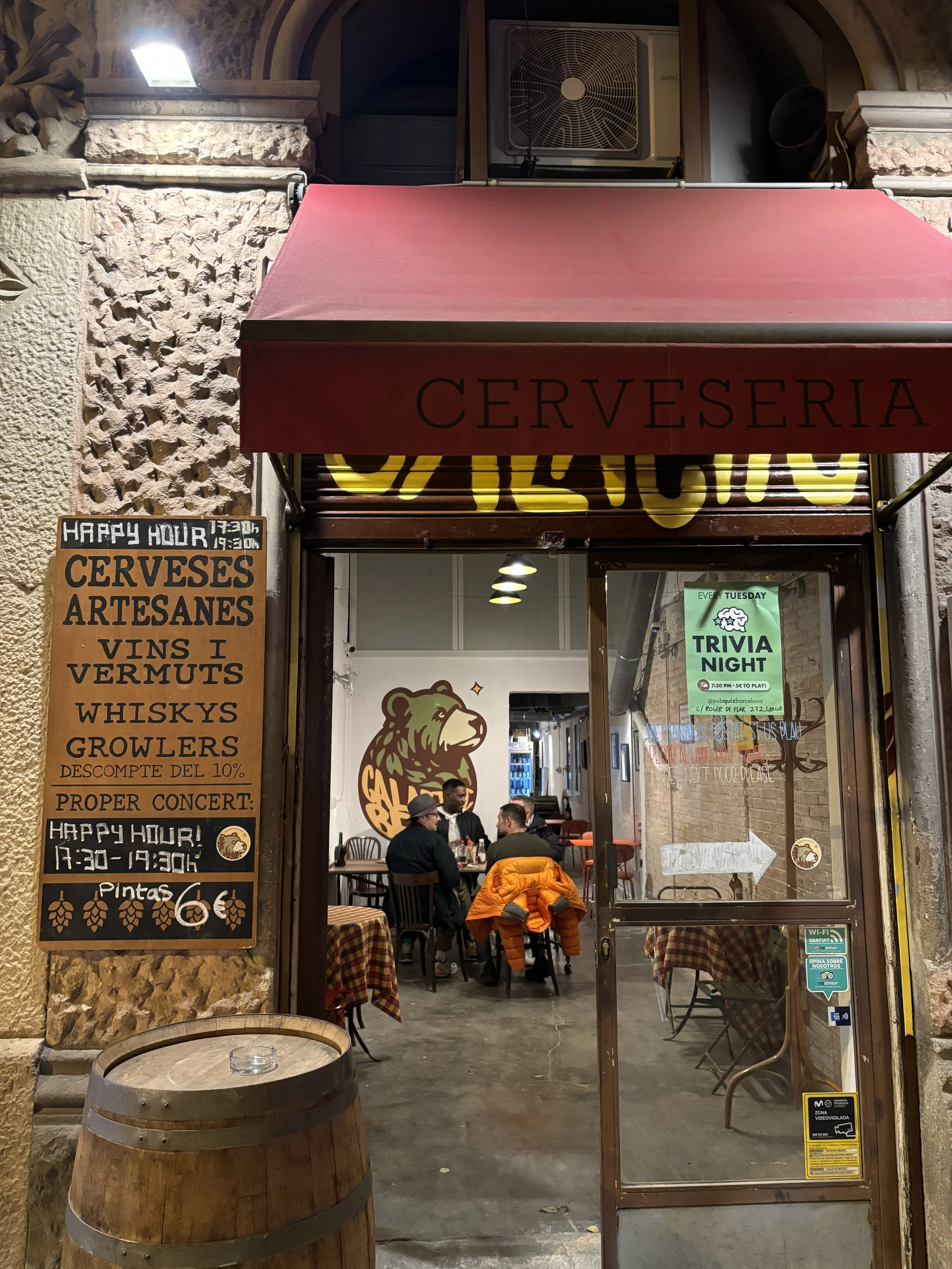 Entrada de un bar o cervecería con un cartel que anuncia promociones y eventos, y vista del interior con varias personas sentadas en mesas de comida, paredes con decoraciones y logotipos de un oso y un león, y un barril de madera en primer plano.