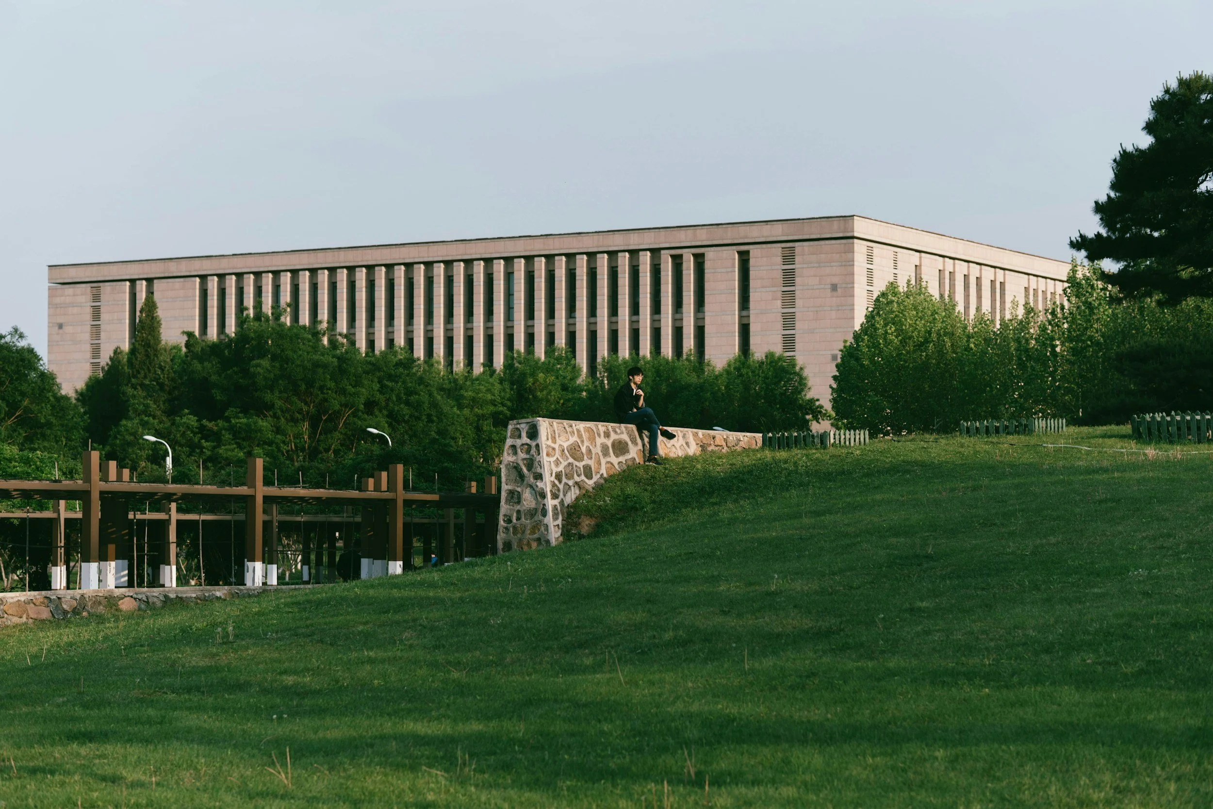 A person sitting on a stone wall in a park with lush green grass and trees, with a large modern building in the background.