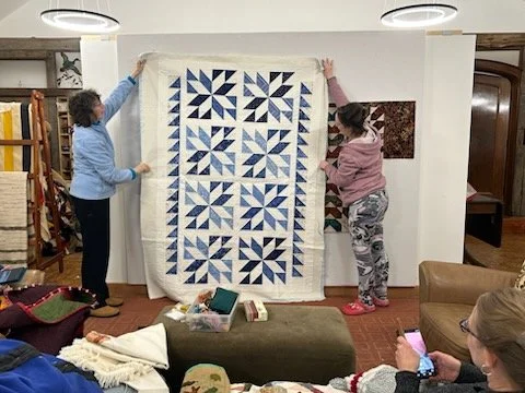Two women hold up a quilt with a blue and white star pattern in a cozy room, with other people sitting and taking photos, surrounded by furniture and decorative items.
