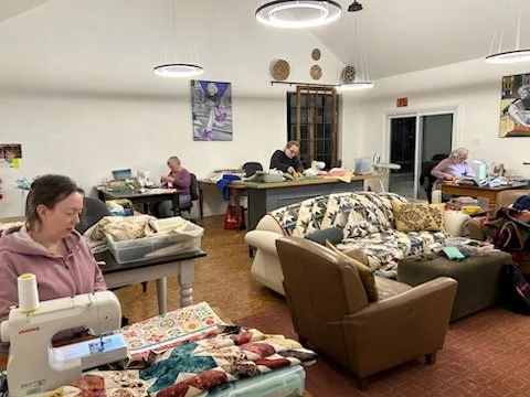 A quilting group in a spacious studio sewing and working on projects, with fabric and supplies on tables and sofas.