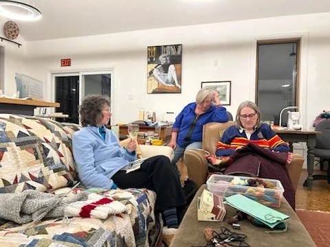 Three women sitting in a cozy living room, engaging in conversation. One woman is sitting on a quilt-covered couch, another on a chair, and the third on the armrest of a chair, with various crafting supplies on a table in front of them.