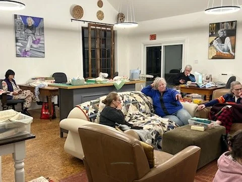 A group of elderly people sitting and talking in a cozy living room with couches and a kitchen area in the background.