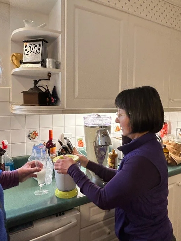 A woman in a purple jacket making a drink using a blender in a kitchen. Another person is holding a champagne glass filled with a drink, and a bottle of wine is on the counter nearby.