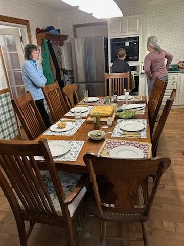 A kitchen with a wooden dining table set for a meal, with six chairs around it. Four people are in the background: three women near the refrigerator and oven, and one woman near a doorway. The table has plates, bowls, glasses, and utensils. The kitch