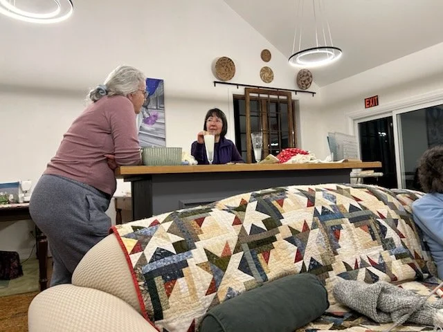 Two women are in conversation at a kitchen counter in a cozy home, with a colorful quilt-covered sofa in the foreground and a large window behind them.