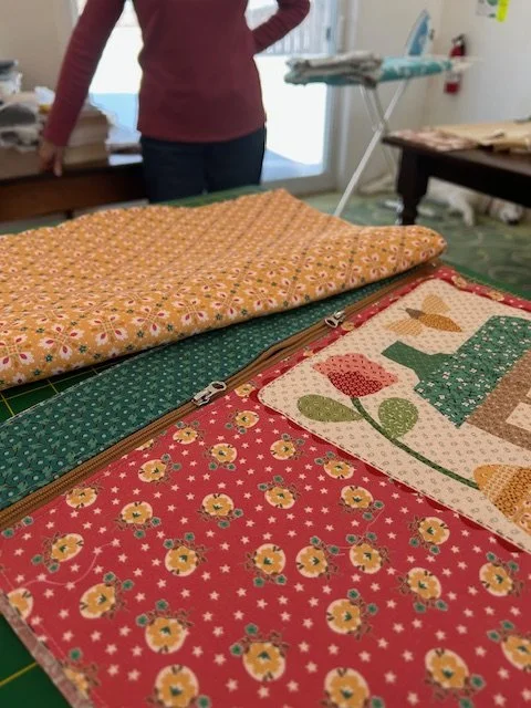 Multiple pieces of fabric with various floral and geometric patterns are laid out on a table in a sewing or craft room, with a person in a red sweater and jeans standing in the background near an ironing board.
