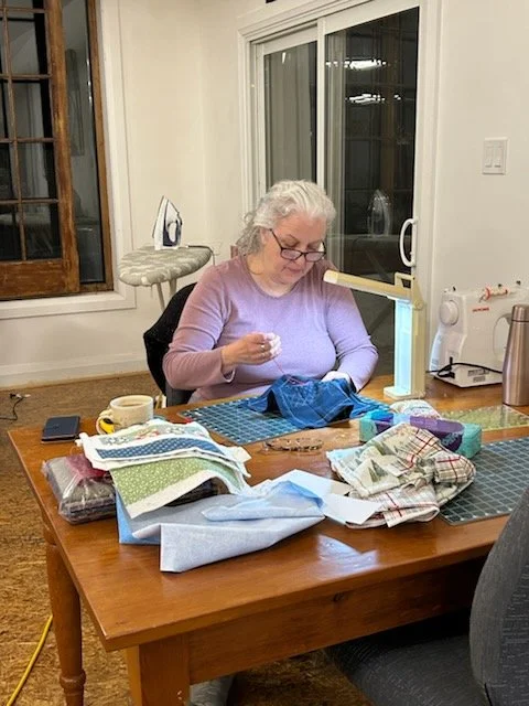 An elderly woman with gray hair and glasses sewing fabric at a wooden table, surrounded by various fabrics and sewing supplies, in a well-lit room with a window and an iron on an ironing board.