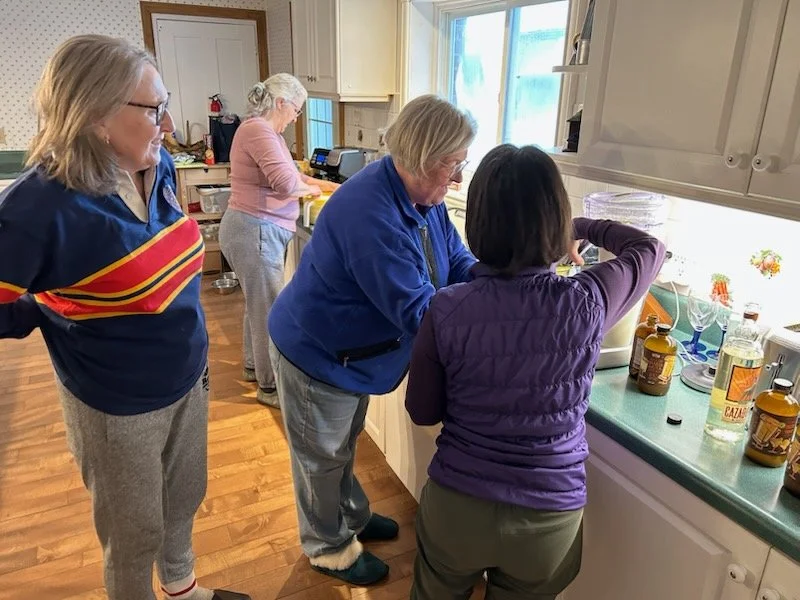 Four women in a kitchen, one woman watching two women preparing food at the countertop, with kitchen cabinets and window in the background.