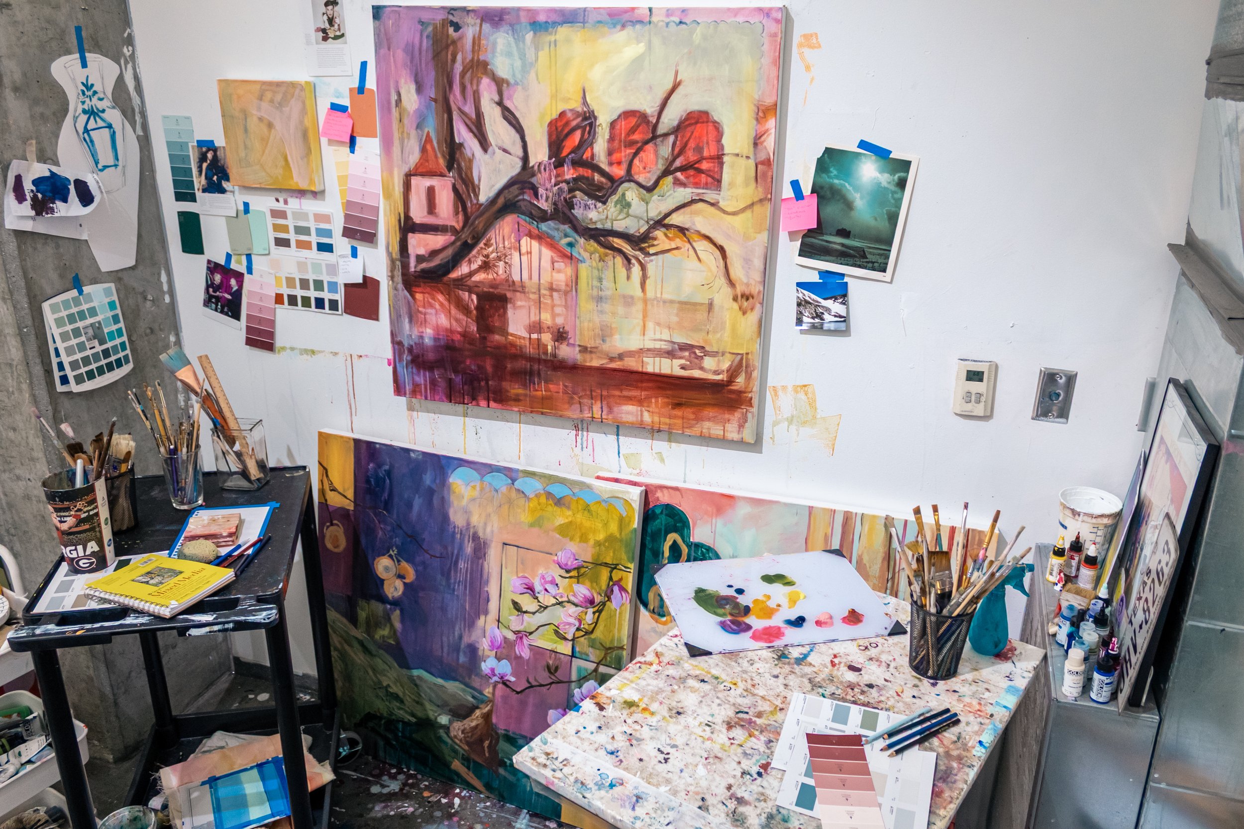 An art studio with paintings, color swatches, and art supplies scattered around. One large painting features a tree with pink and red foliage, with dripping paint effect. Smaller paintings include a scene with flowers and butterflies. Various brushes
