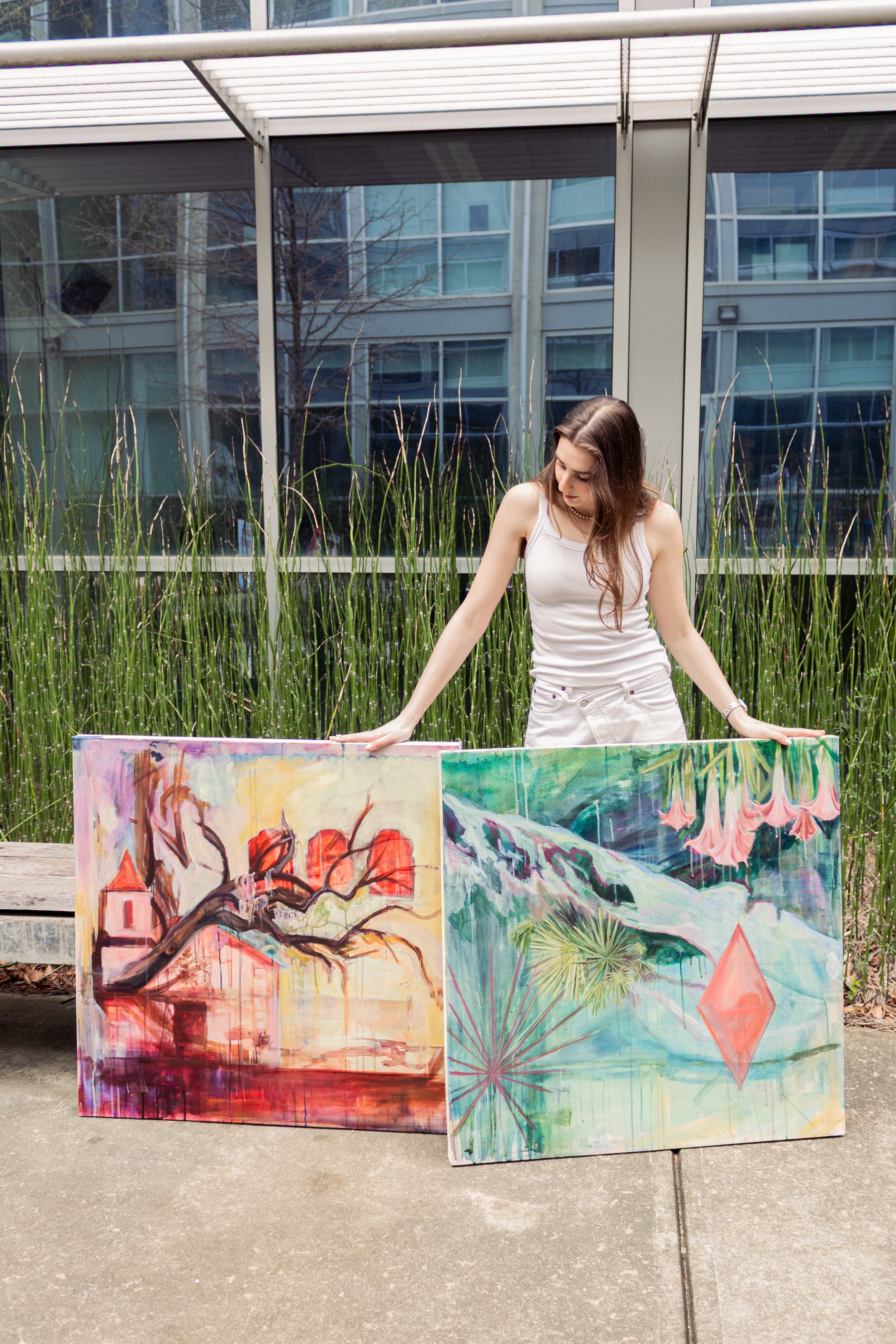 A woman with long brown hair wears a white sleeveless top and white shorts, standing outdoors behind two colorful paintings on canvas, with a glass building and greenery behind her.