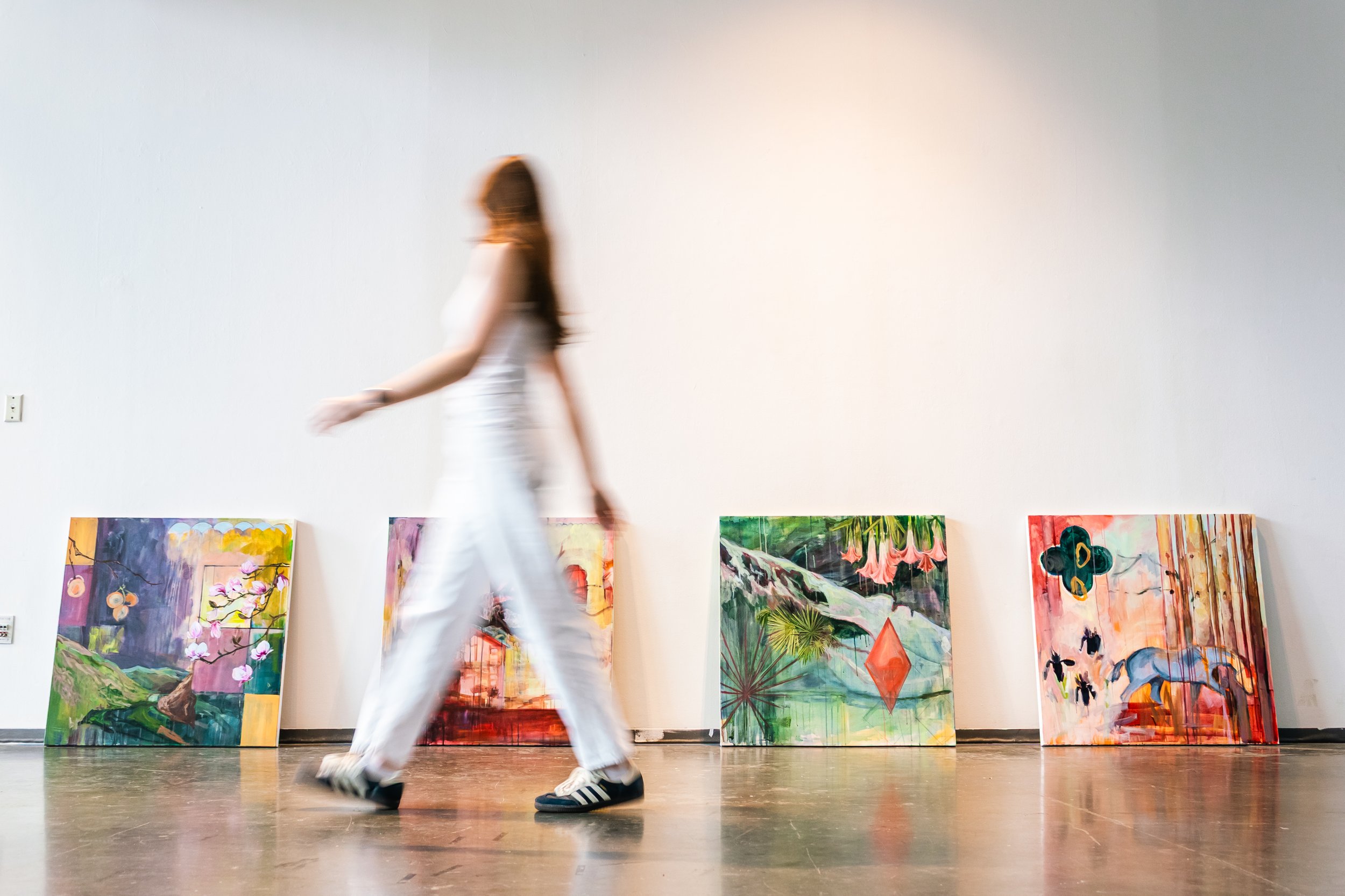A woman with long hair walking past a row of colorful paintings displayed against a white wall in an art gallery.