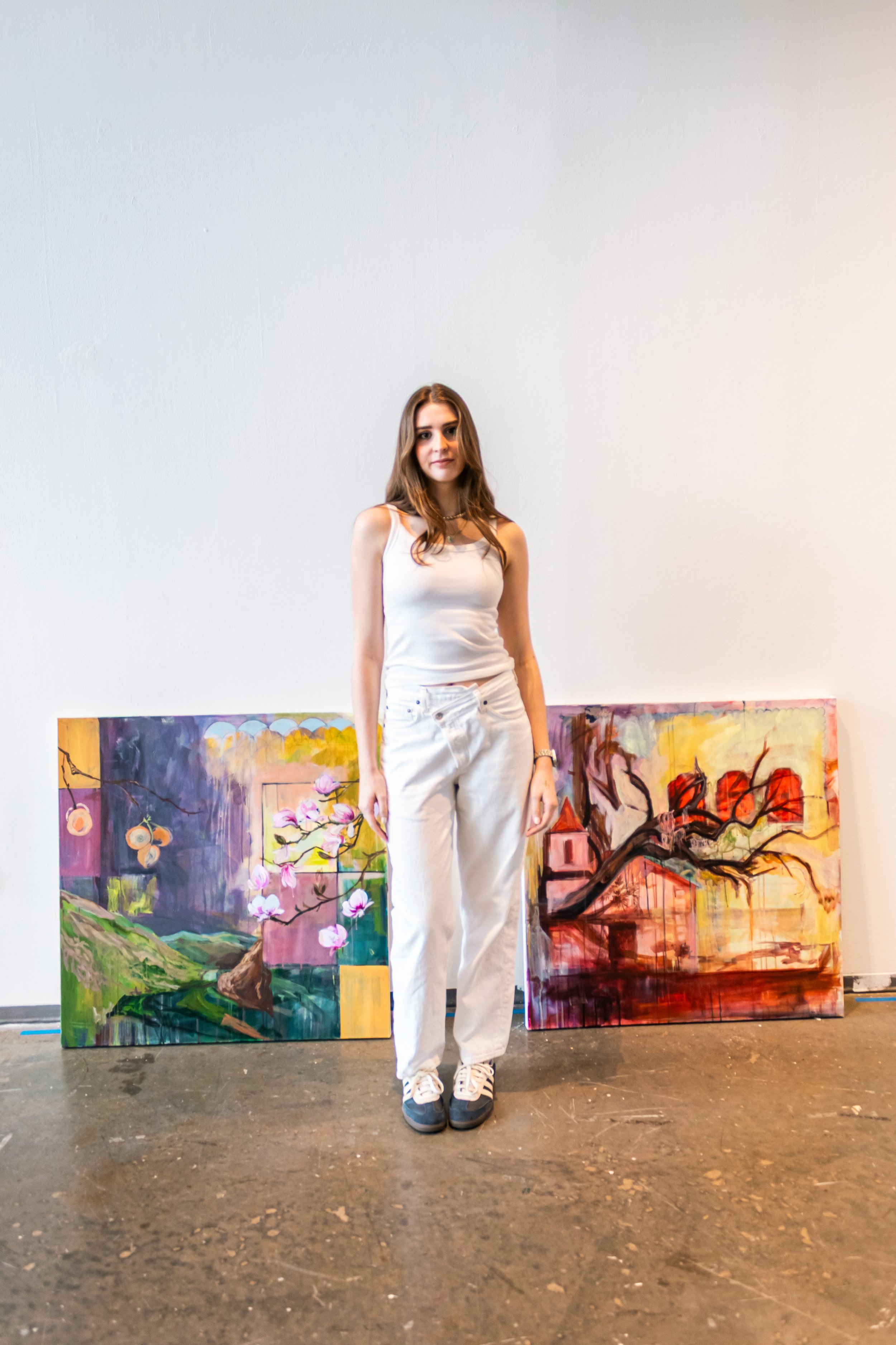 Young woman standing in front of two colorful paintings, wearing a white tank top and white pants.