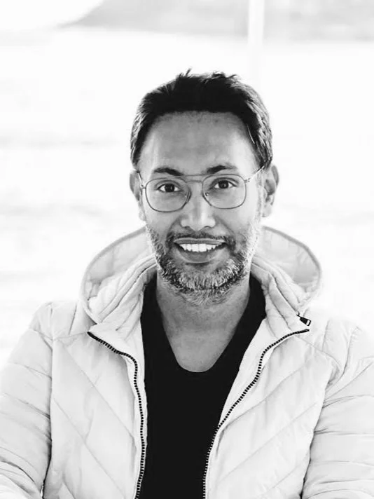 Person wearing glasses with curly hair and a beard, dressed in a dark sweater, looking to the side.