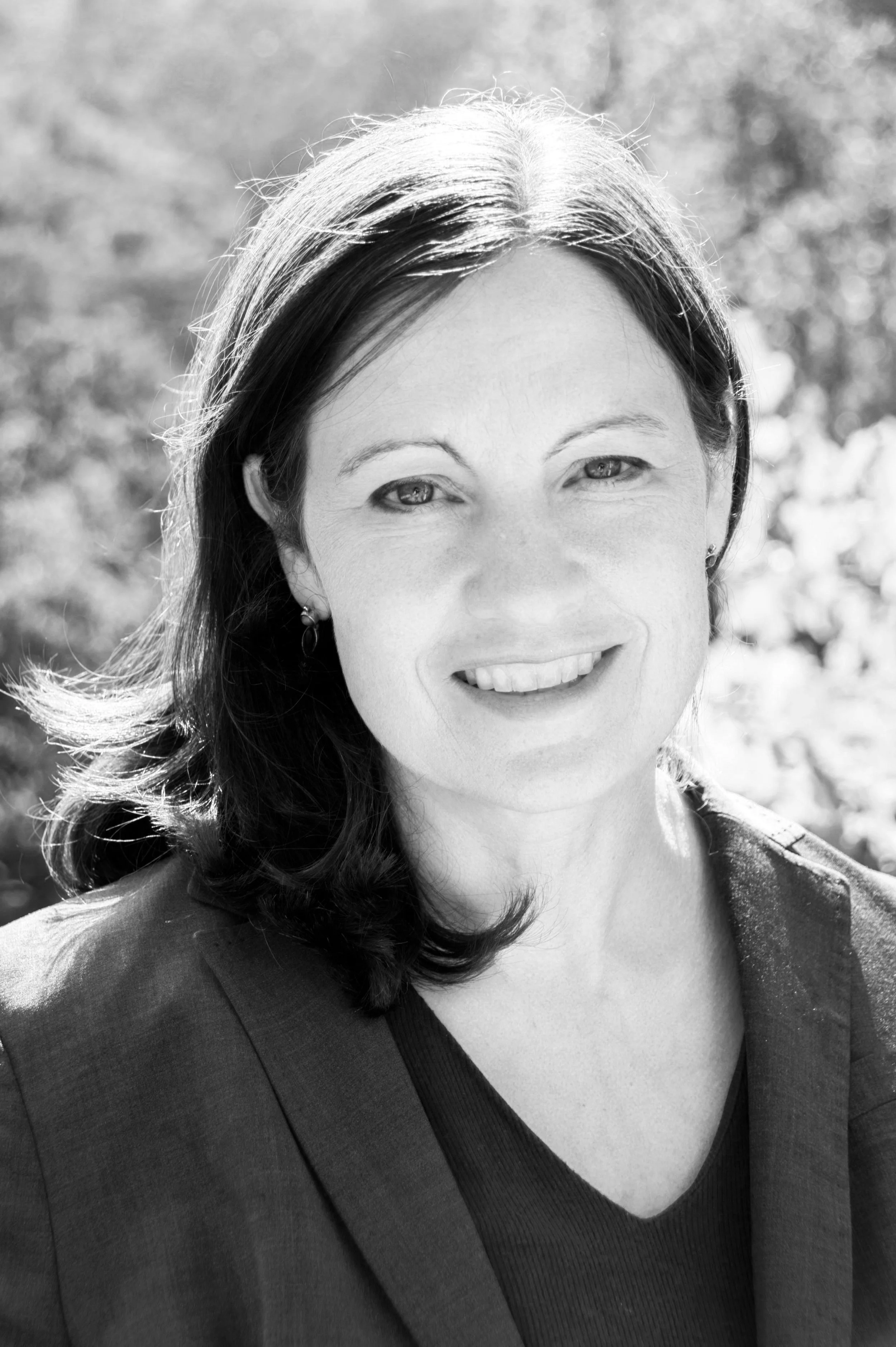 Black and white portrait of a smiling person with medium-length hair, wearing a blazer, standing outdoors.